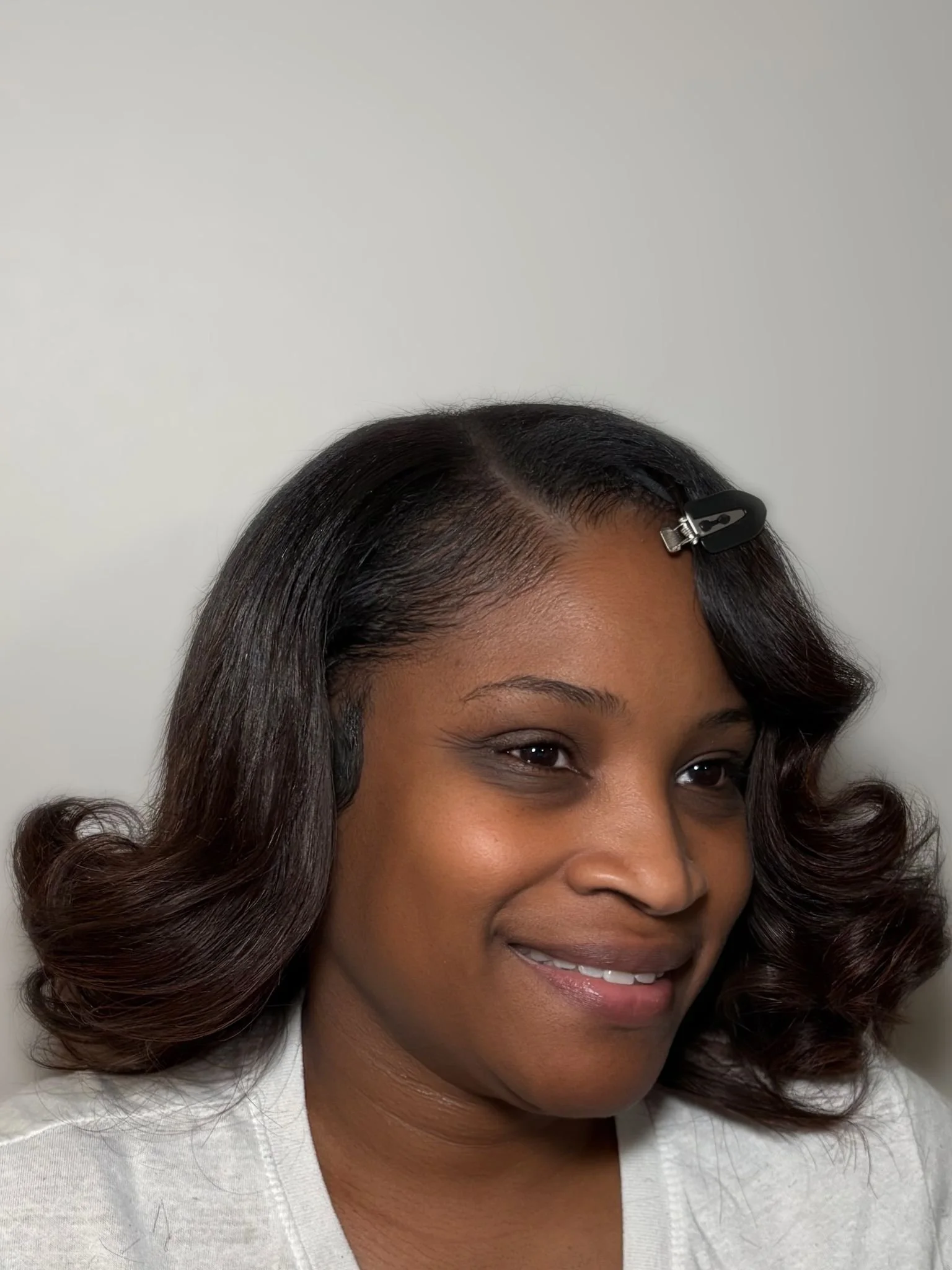 Close-up of a woman with dark, styled hair held in place by a black hair clip, smiling slightly, wearing a white shirt, against a light-colored background.