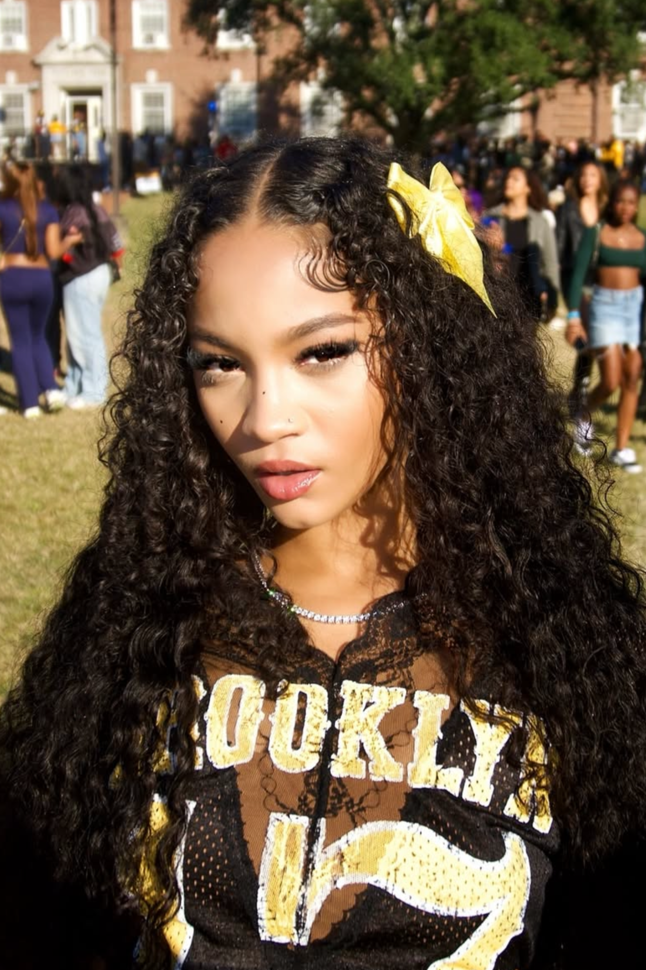 Close-up of a young woman with long, curly hair, wearing a yellow bow in her hair, and a black and yellow top with the word 'Hook' visible, outdoors at a daytime event with a crowd and a brick building in the background.