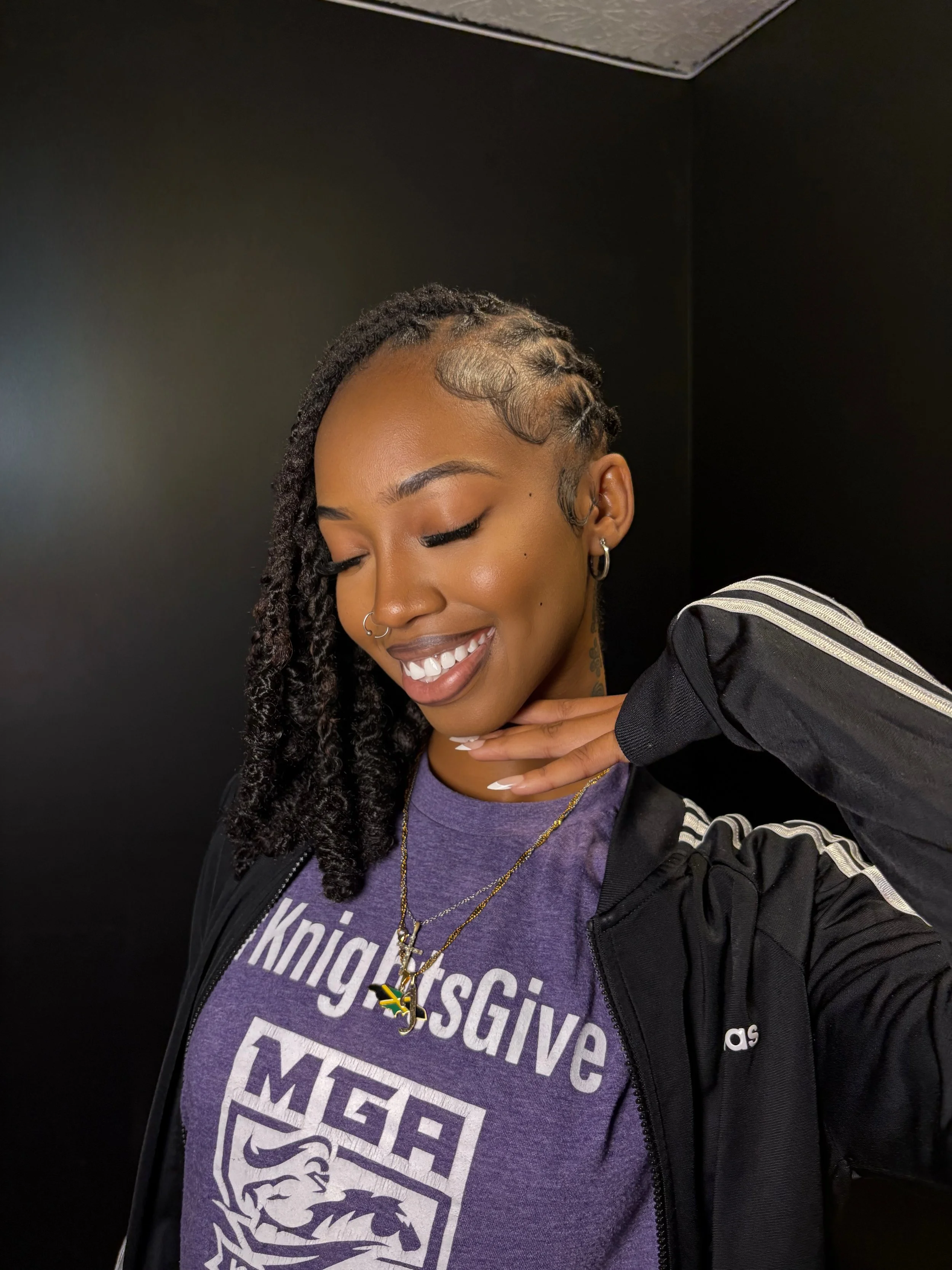 Smiling woman with dreadlocks, wearing a purple T-shirt with text, a black jacket, and jewelry, against a black background.