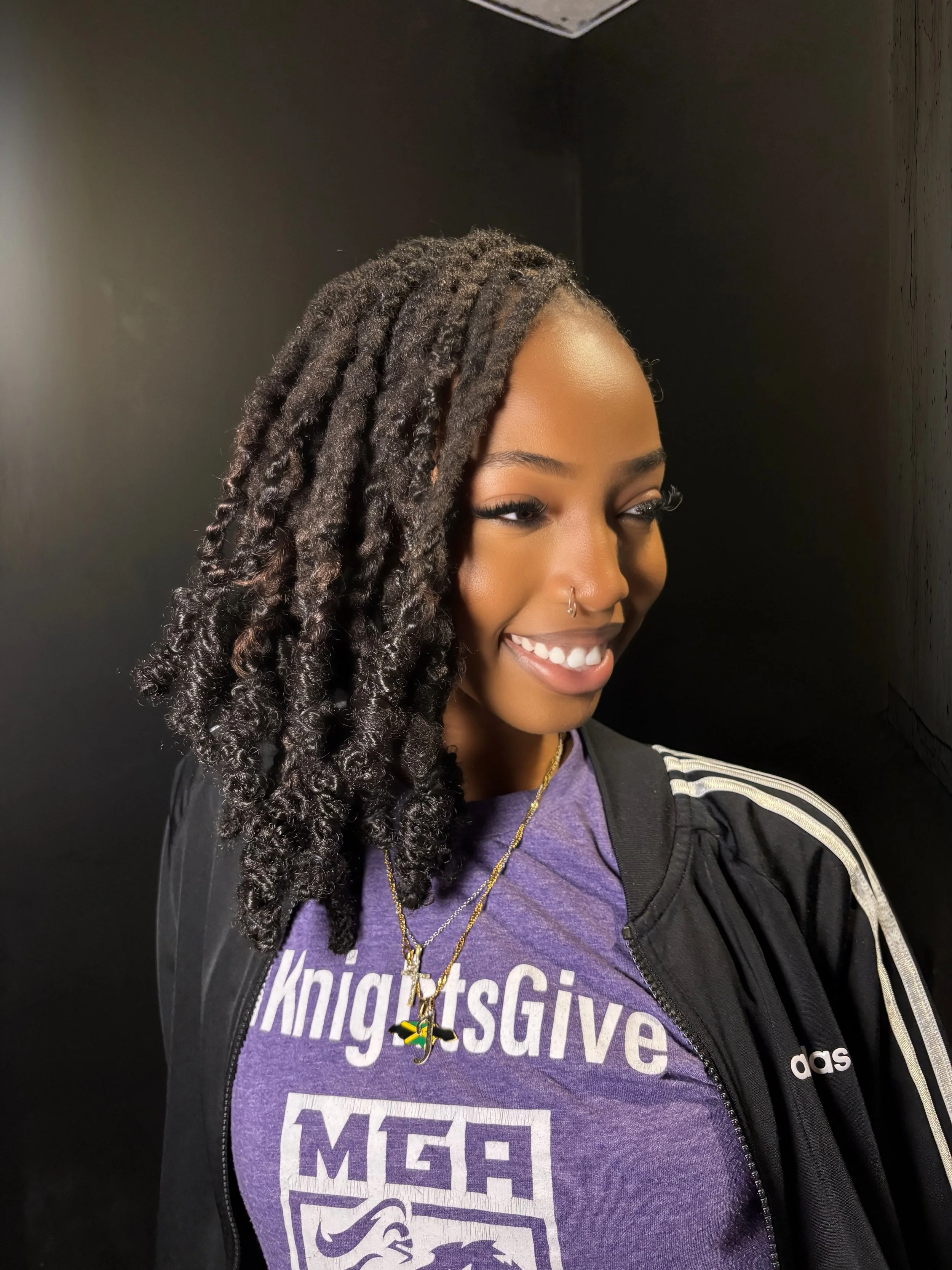 Close-up of a young woman with dark, curly, dreadlock hairstyle, smiling, wearing a purple T-shirt with white text, a black Adidas jacket, and gold necklaces, against a black background.