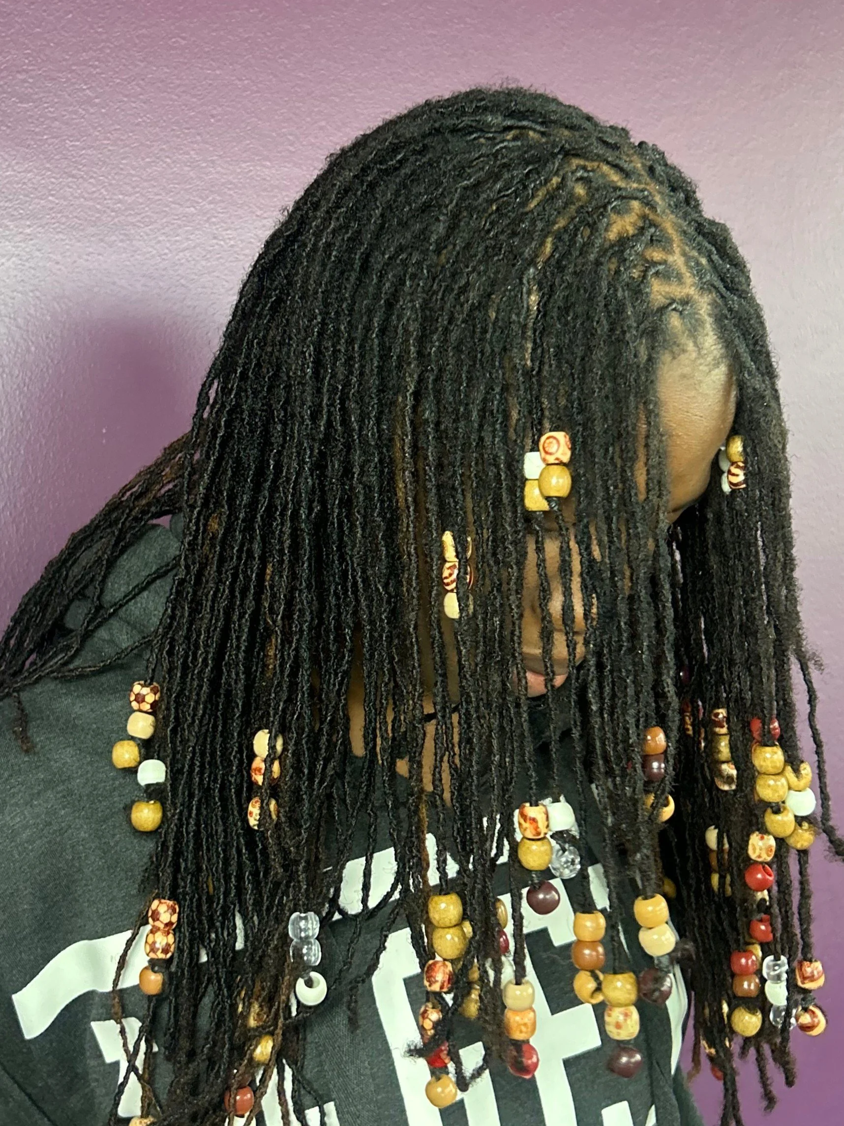 Close-up of a person with long black dreadlocks decorated with various wooden and bead accessories, wearing a black shirt with partially visible white text, against a purple wall background.