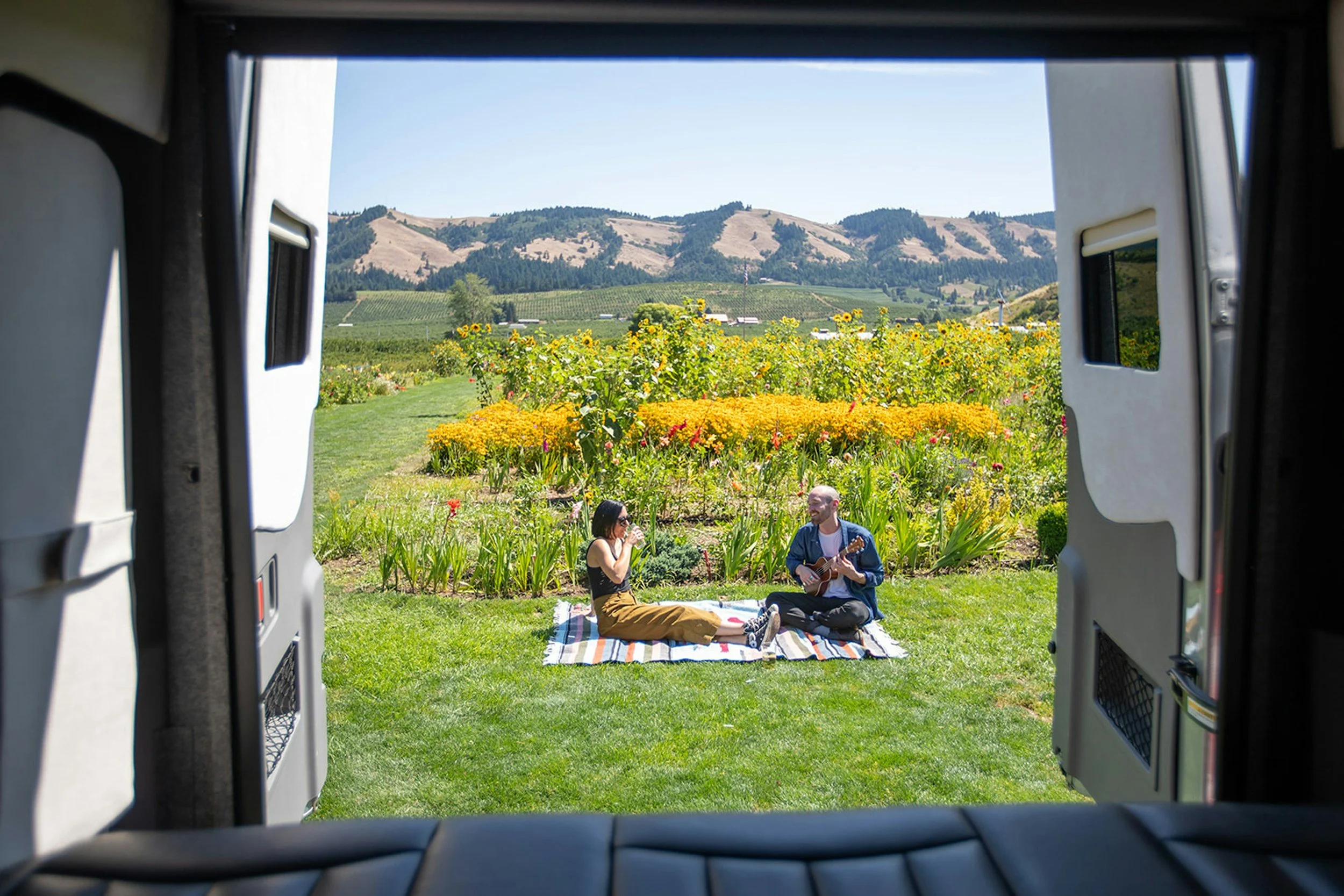 Two people having a picnic and playing guitar on a colorful blanket in a garden with yellow and orange flowers, viewed through the back of a van, with hills and a clear sky in the background.