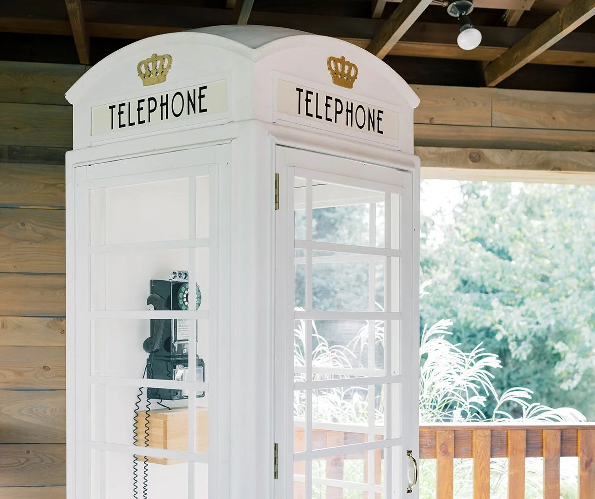 White wooden phone booth with a vintage black rotary phone inside, labeled 'TELEPHONE' with a gold crown above, located on a wooden porch with a view of green trees outside.