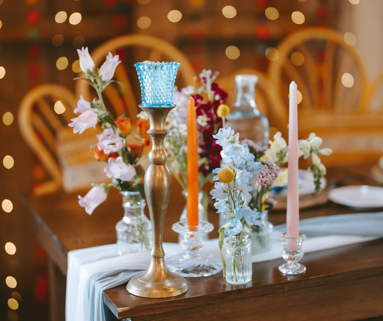 Decorative table setting with colorful flowers, tall candlesticks, and a white table runner, set in a warmly lit room with blurred string lights in the background.