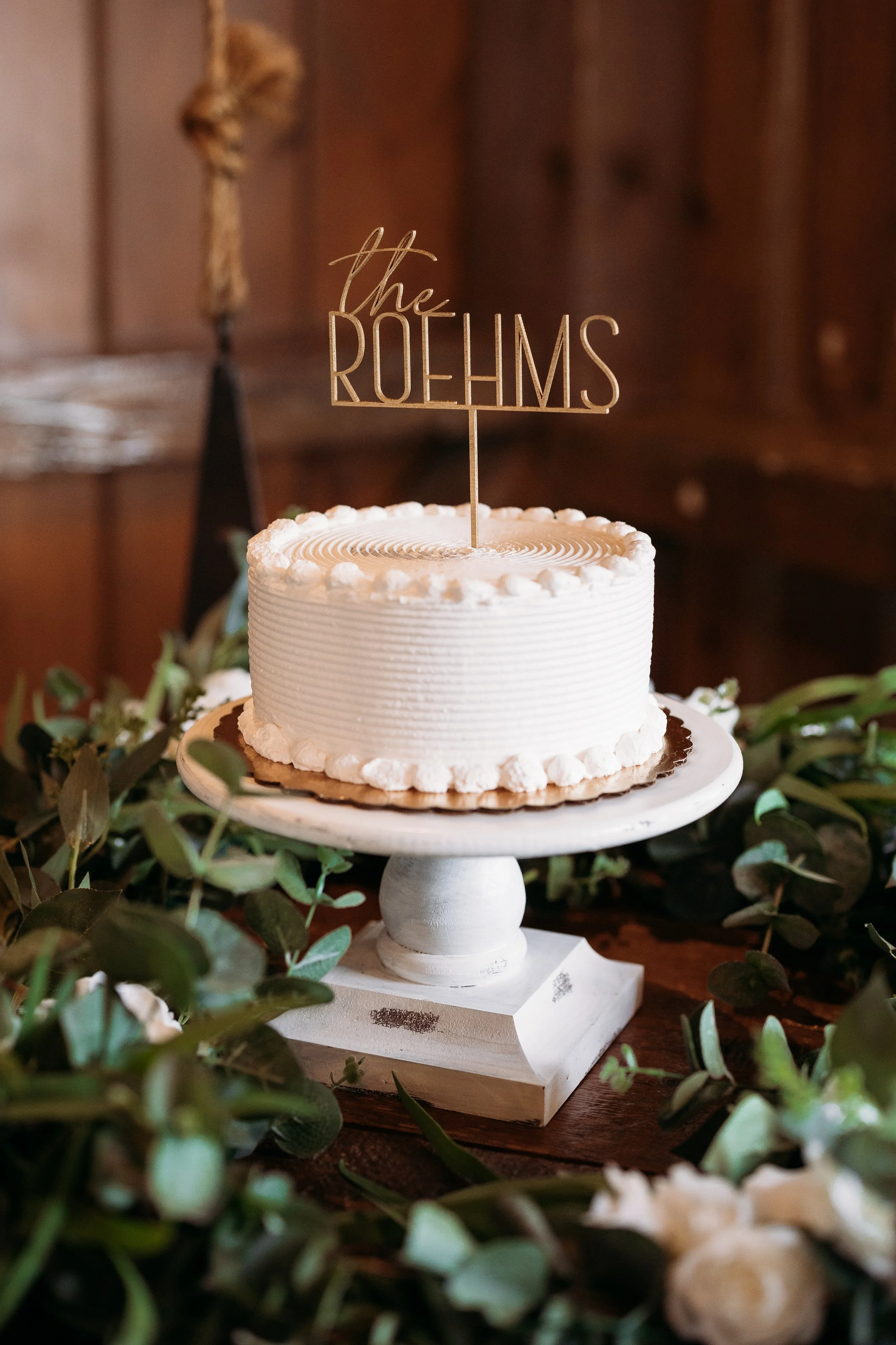A white frosted birthday cake with a gold 'the ROEHMS' cake topper, decorated with piped white icing, placed on a white cake stand, surrounded by greenery and small white flowers, with a blurred wooden background.