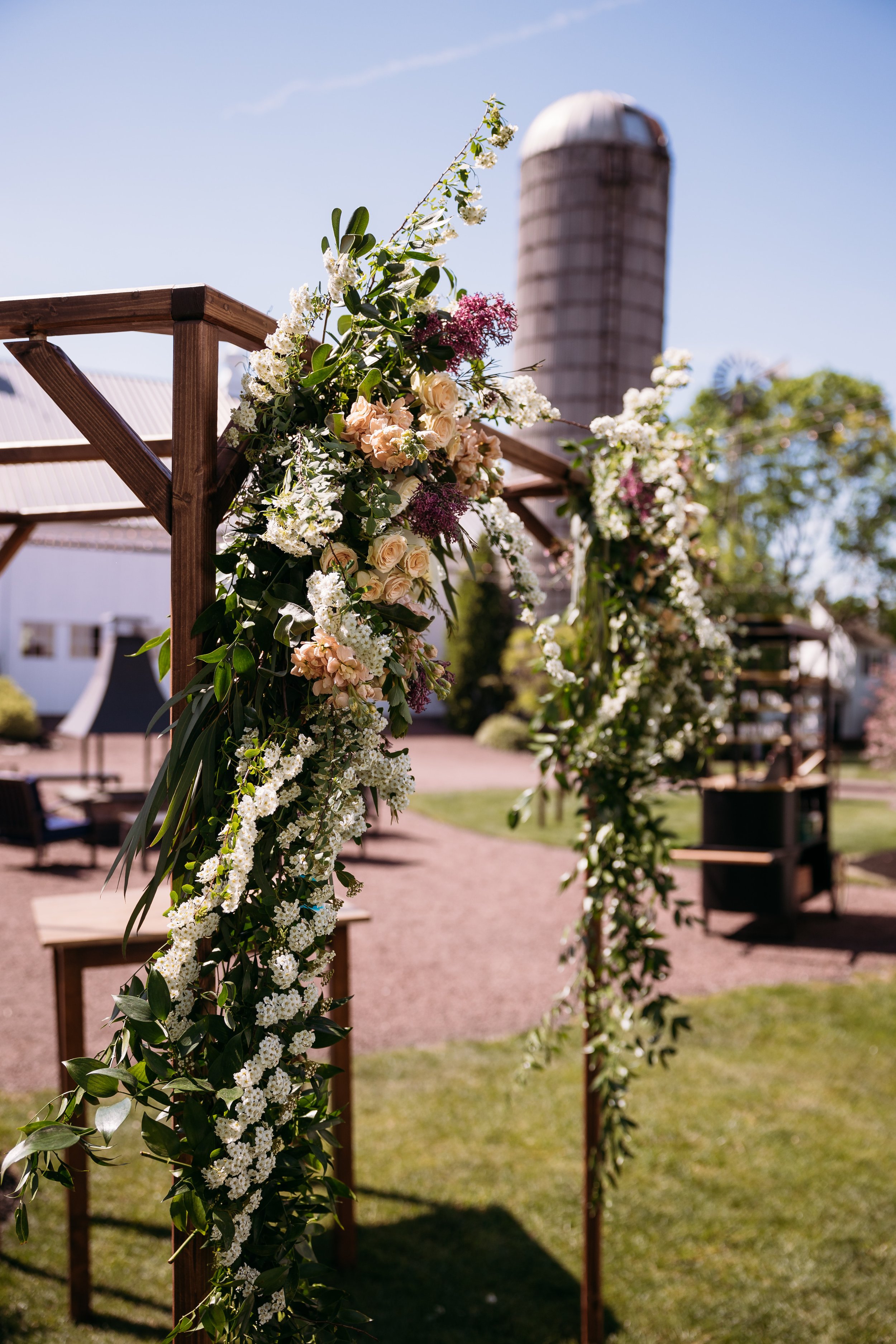 Close-up of a floral wedding arch with white, pink, and purple flowers, set outdoors under a clear blue sky with a rustic barn, trees, and seating in the background.