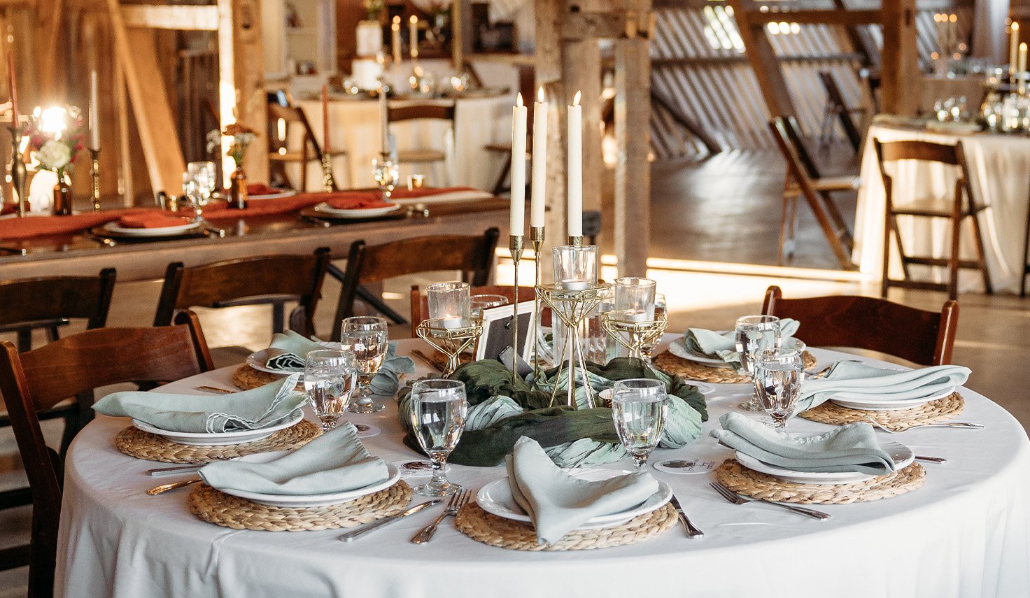 Round dining table set for a meal with white tablecloth, woven placemats, folded white napkins, wine glasses, and a centerpiece with candles and greenery in a rustic wooden setting.