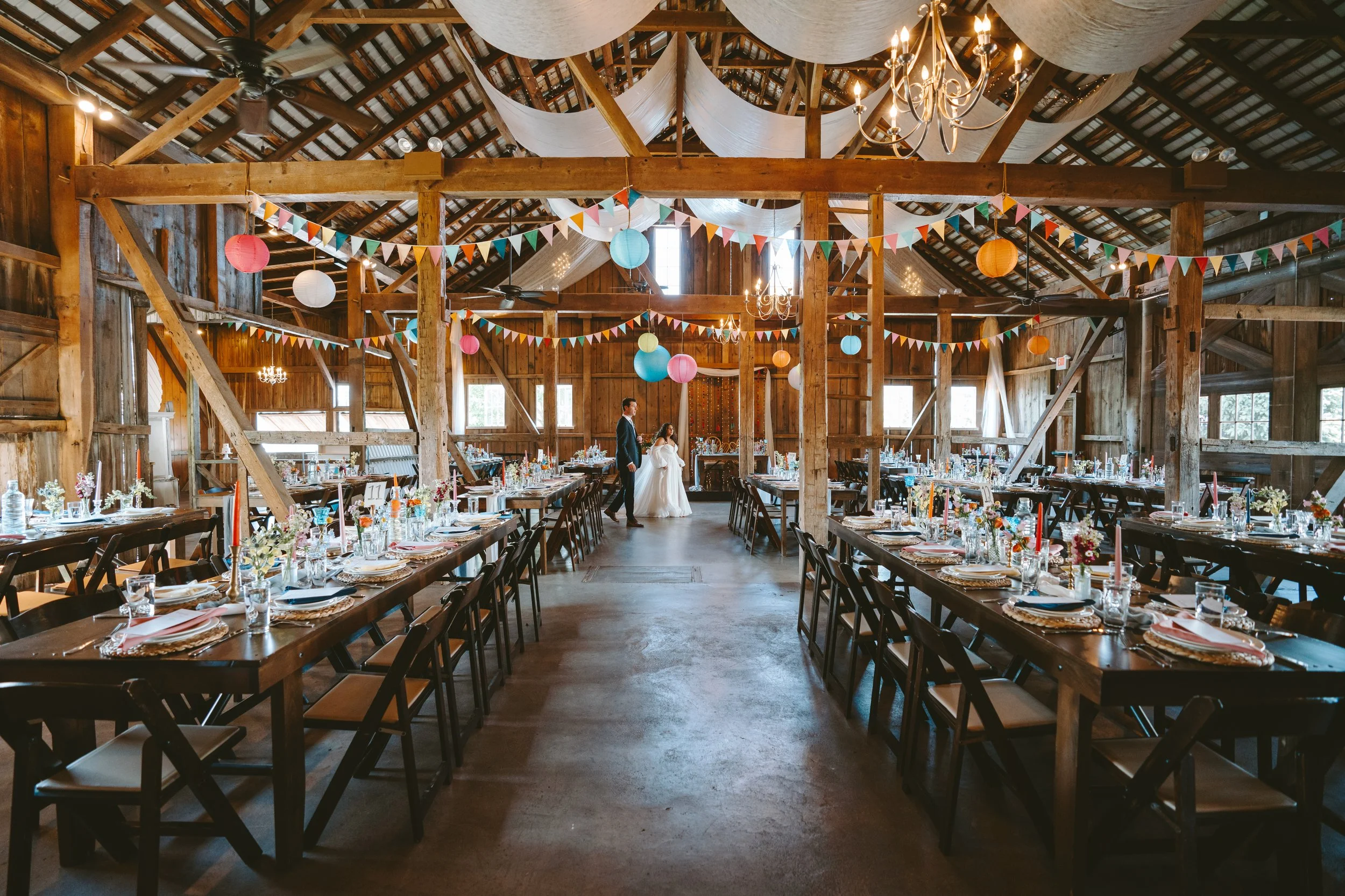 Interior of a rustic barn decorated for a wedding reception with colorful bunting and paper lanterns, featuring long banquet tables set with floral centerpieces and place settings, and a bride and groom standing together at the far end.