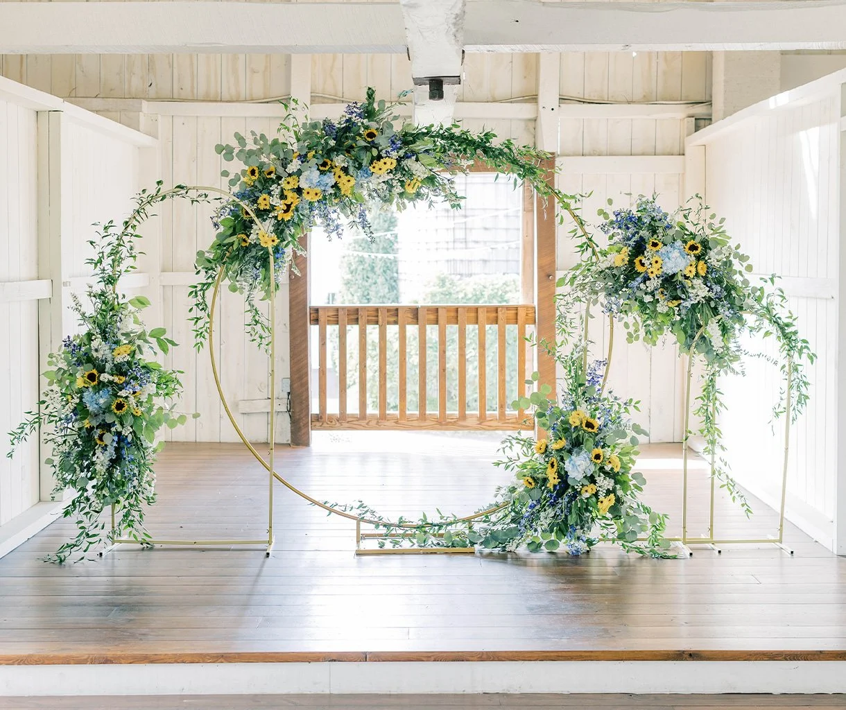 A floral wedding arch with yellow and blue flowers and green foliage in a white wooden barn setting.