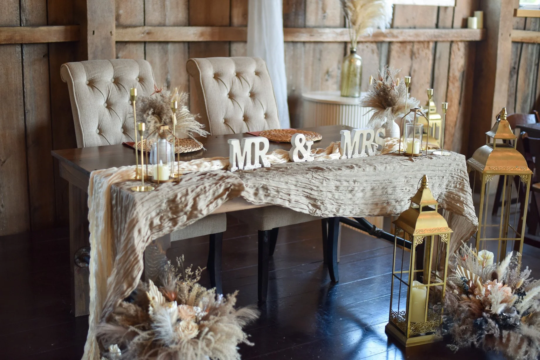 A rustic wedding reception table setup with beige tufted chairs, a beige tablecloth, decorative 'MR & MRS' signs, and gold accents. The table features floral arrangements, candles, and decorative gold lanterns in a warmly lit wooden room.