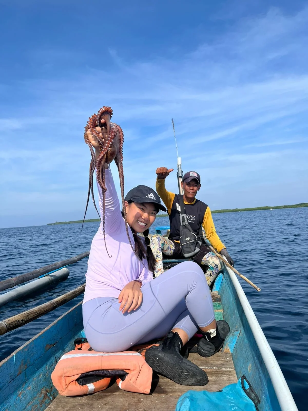 Two people in a small boat on the water, holding an octopus, with one person smiling at the camera and the other giving a thumbs-up. Clear blue sky and distant shoreline in the background.