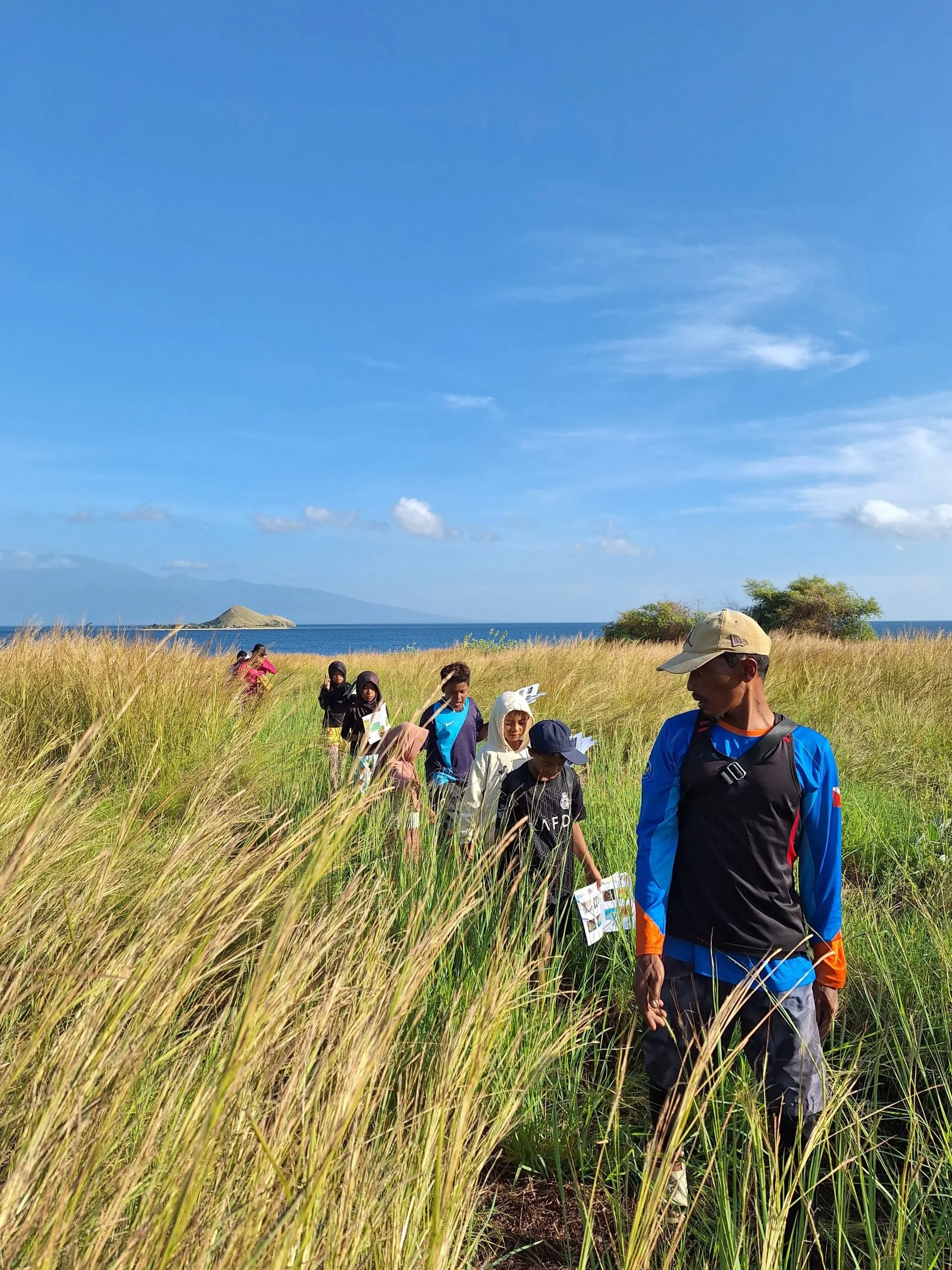 A group of people walking through tall grass on a trail near a body of water, with mountains in the distance and a blue sky overhead.