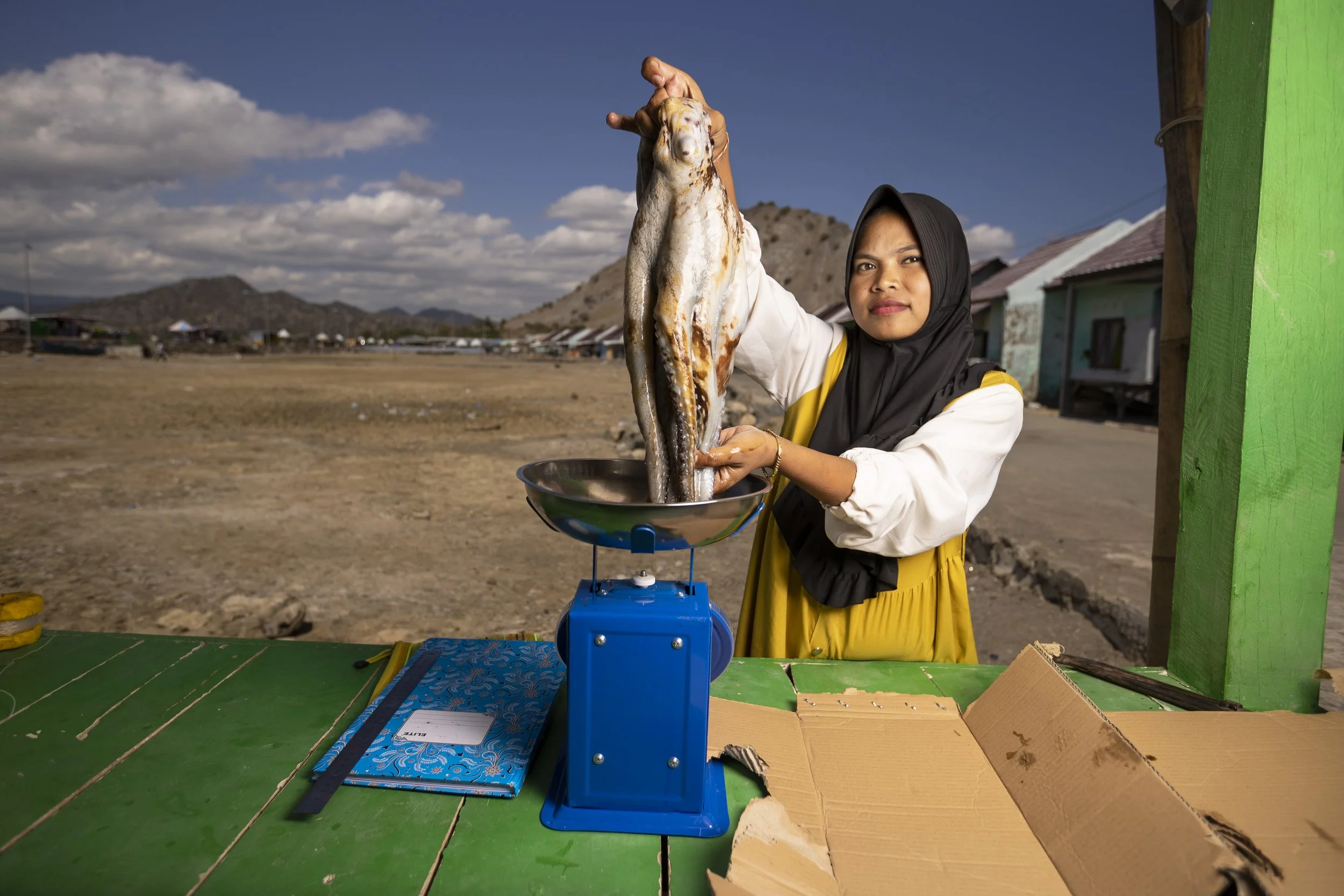 A woman wearing a black hijab and a yellow and white shirt holding a large octopus over a blue scale. The scene is outdoors with mountains, houses, and a partly cloudy sky in the background.
