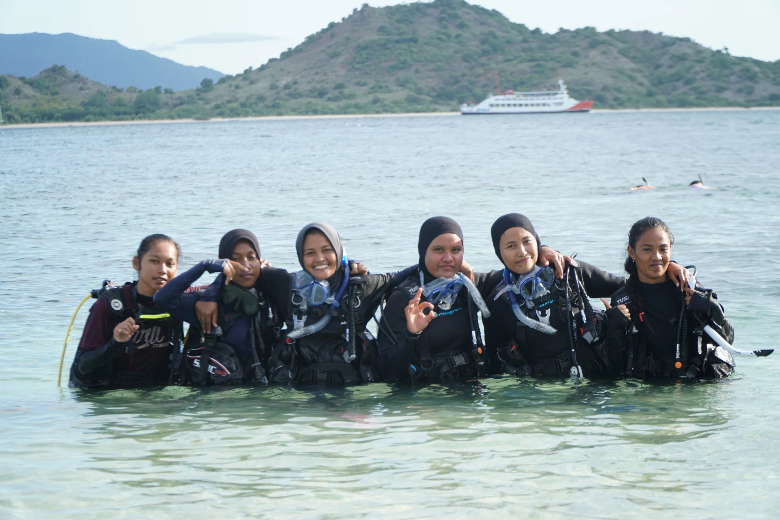 Group of six women in scuba gear standing in water at the beach with a mountain and boat in the background.