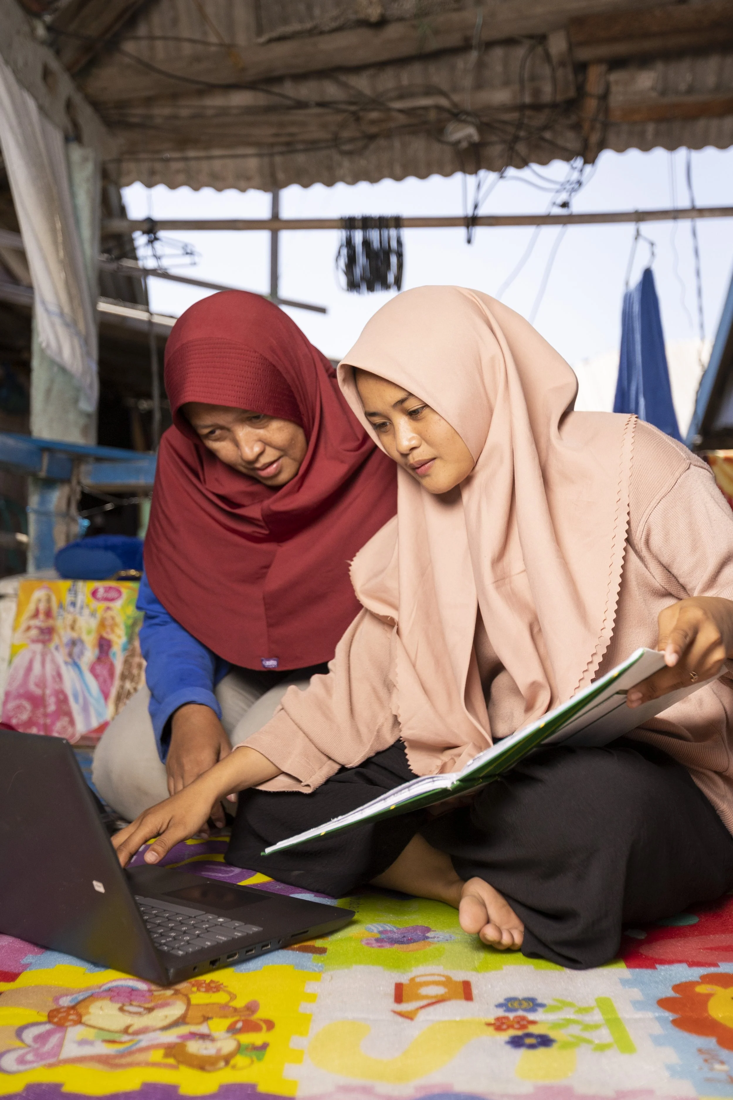 Two women wearing hijabs, one red and one beige, sitting on a colorful children's playmat, working on a laptop and book in a simple, open-air space with a wooden roof and hanging clothes.