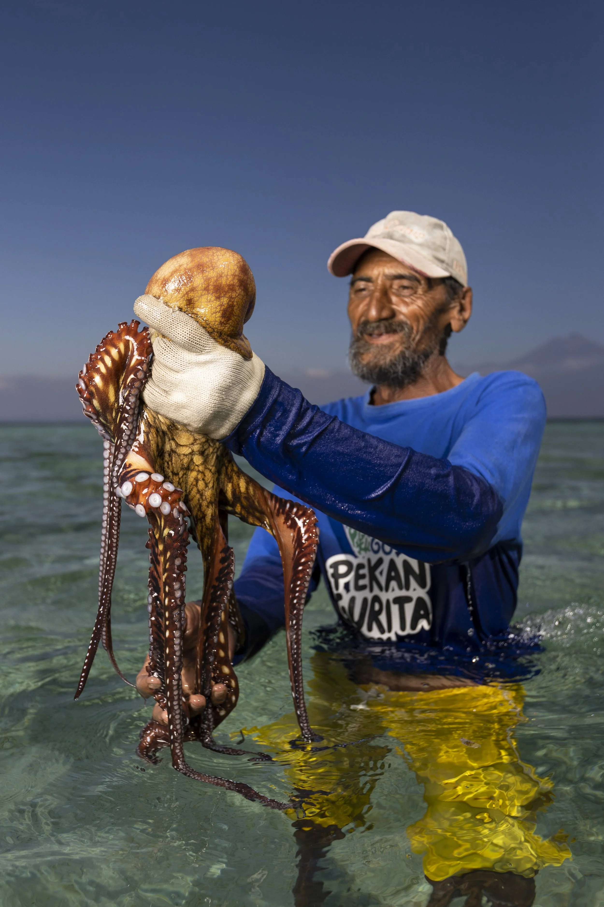 A man in a pink cap and blue shirt holding a large octopus in clear water with a blue sky background.