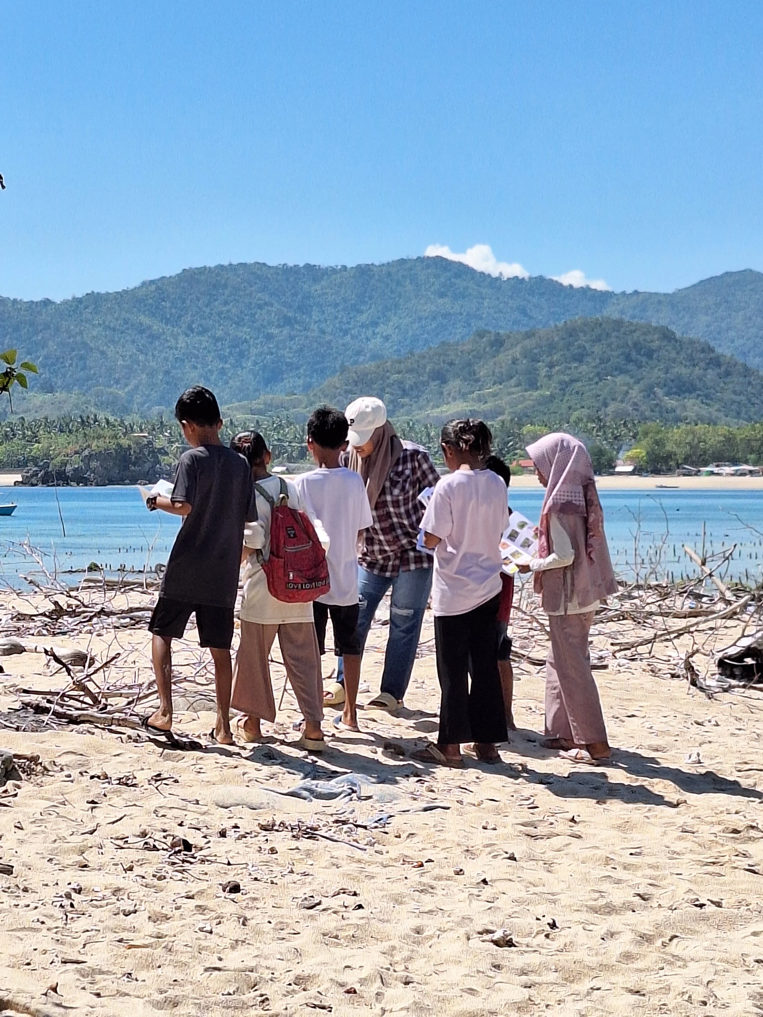 A group of people standing on a sandy beach with mountains and clear blue sky in the background, some reading papers and talking.