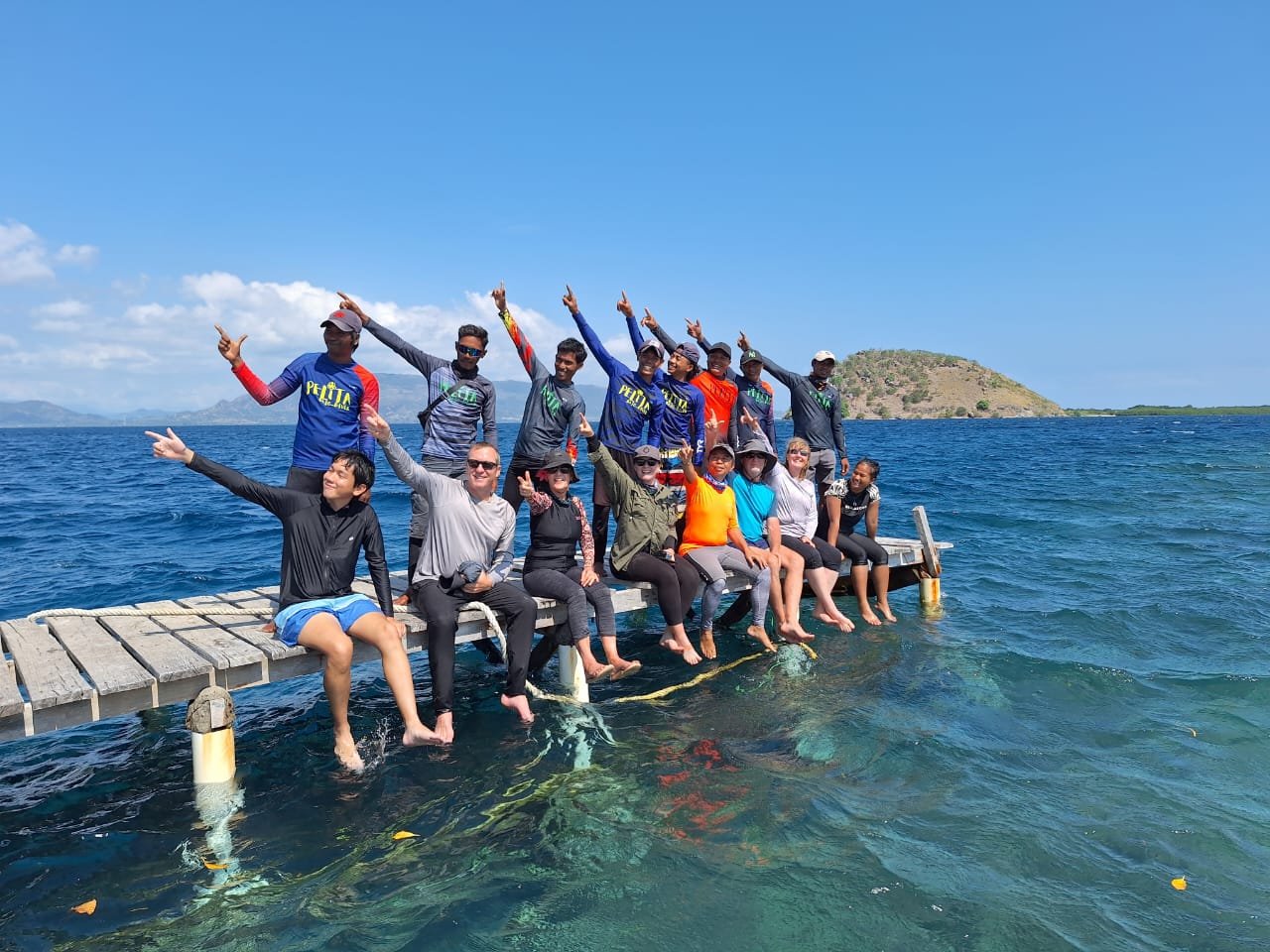 A group of people sitting and standing on a small dock over the water, pointing upwards and smiling, with a bright blue sky and distant land in the background.