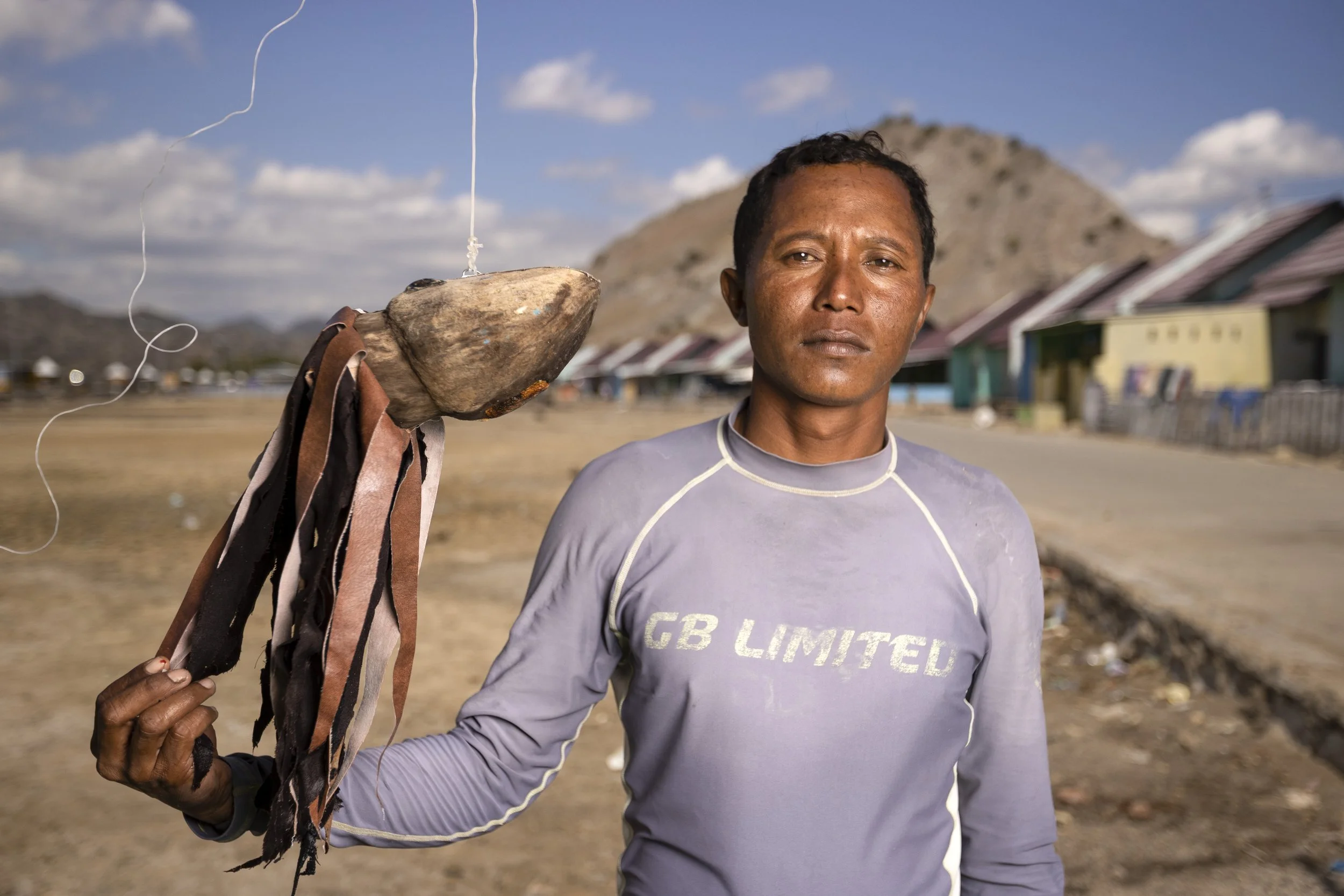 A man holding a rock with strips of leather or fabric tied to it, standing outdoors in a rural area with mountains and houses in the background.