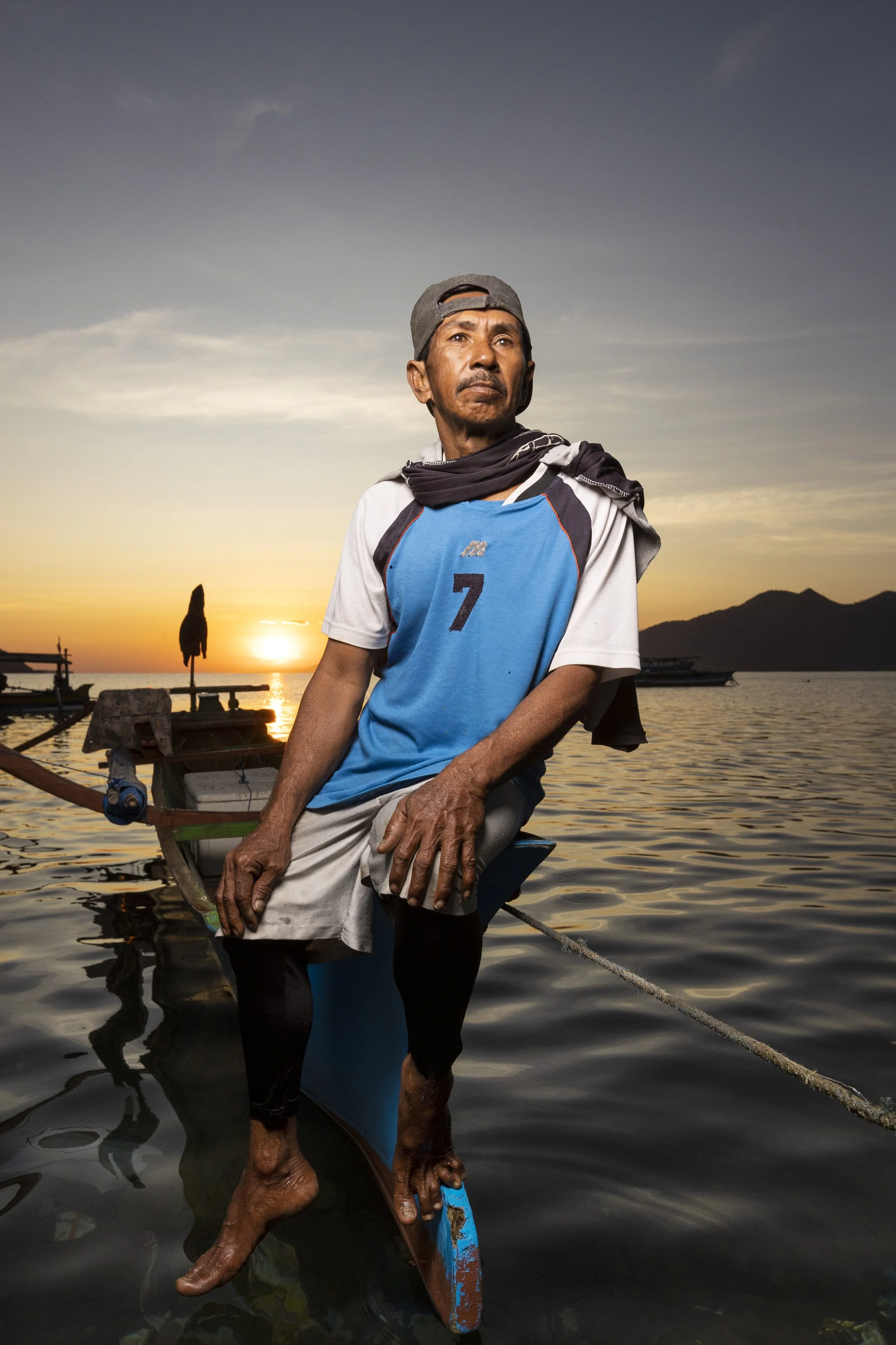 Man standing in shallow water during sunset, wearing a blue sports jersey with the number 7, a gray cap, and a black scarf, with boats and mountains in the background.
