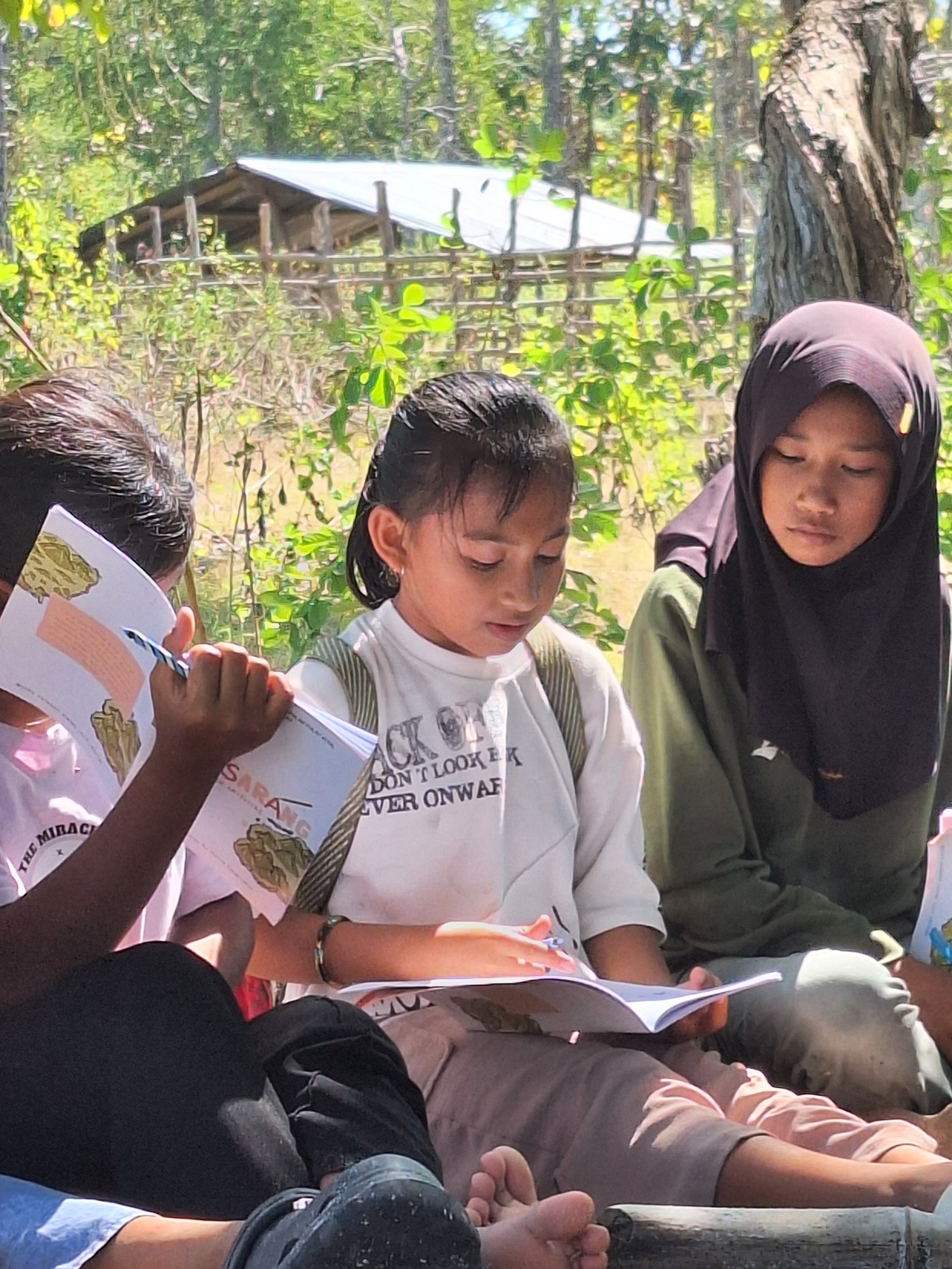 Three children sitting outdoors, reading or looking at papers. One girl at the center is focused on what she is reading, wearing a white shirt, and has short dark hair. To her right is a girl wearing a black hijab and a green jacket, also looking at papers. The arm and hand of a child holding a booklet with an illustrated map are visible on the left side of the image. The background features trees and an elevated wooden structure with a tin roof.