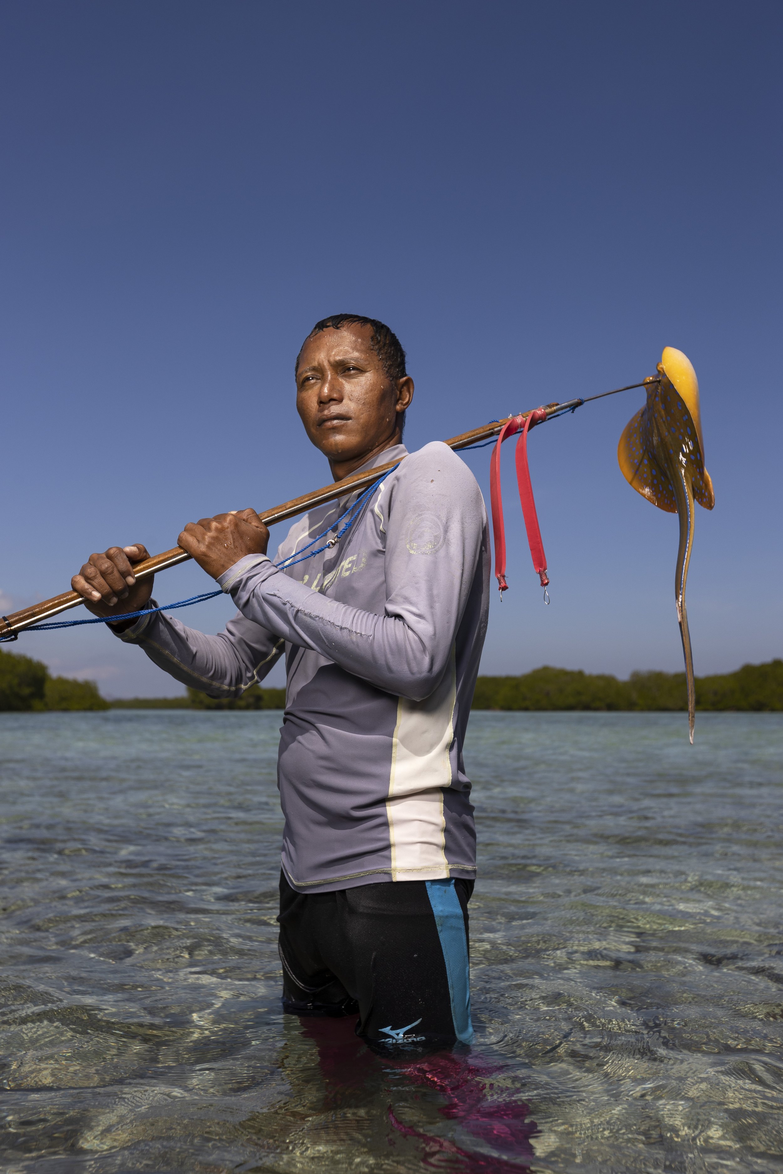 A man holding a fishing pole with a fish on the line, standing waist-deep in water with a clear blue sky above and trees in the background.