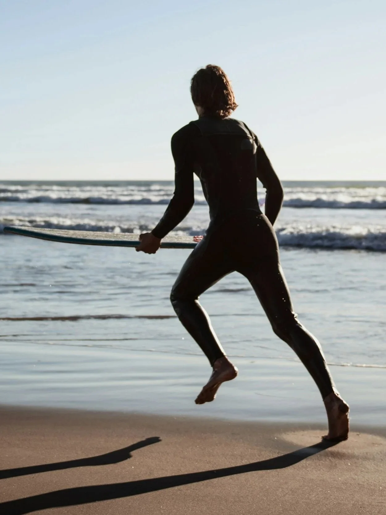 Person in a wetsuit running on the beach with a surfboard, near the ocean during sunset.