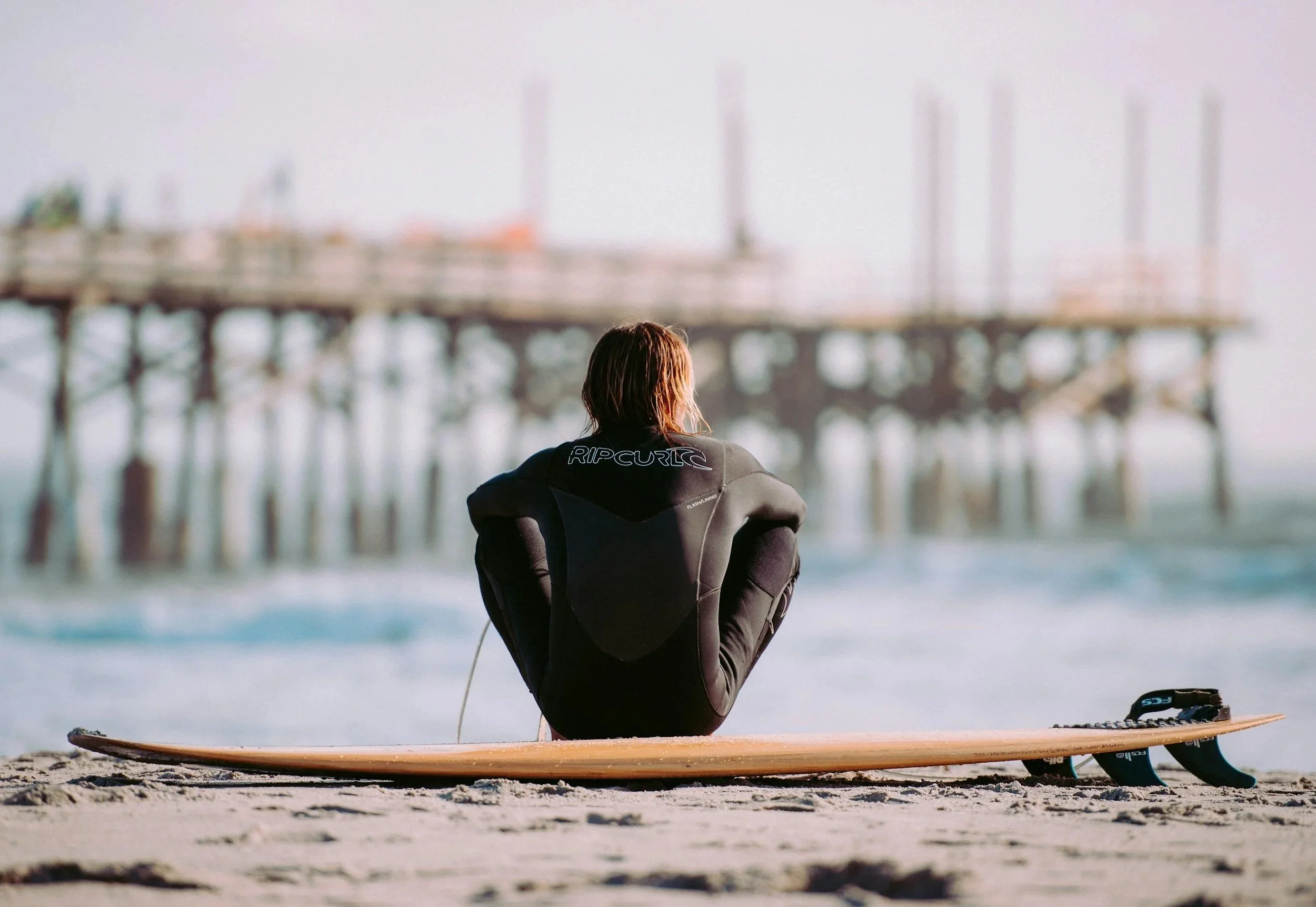 A person sitting on a surfboard at the beach, watching an offshore pier with boats in the distance.
