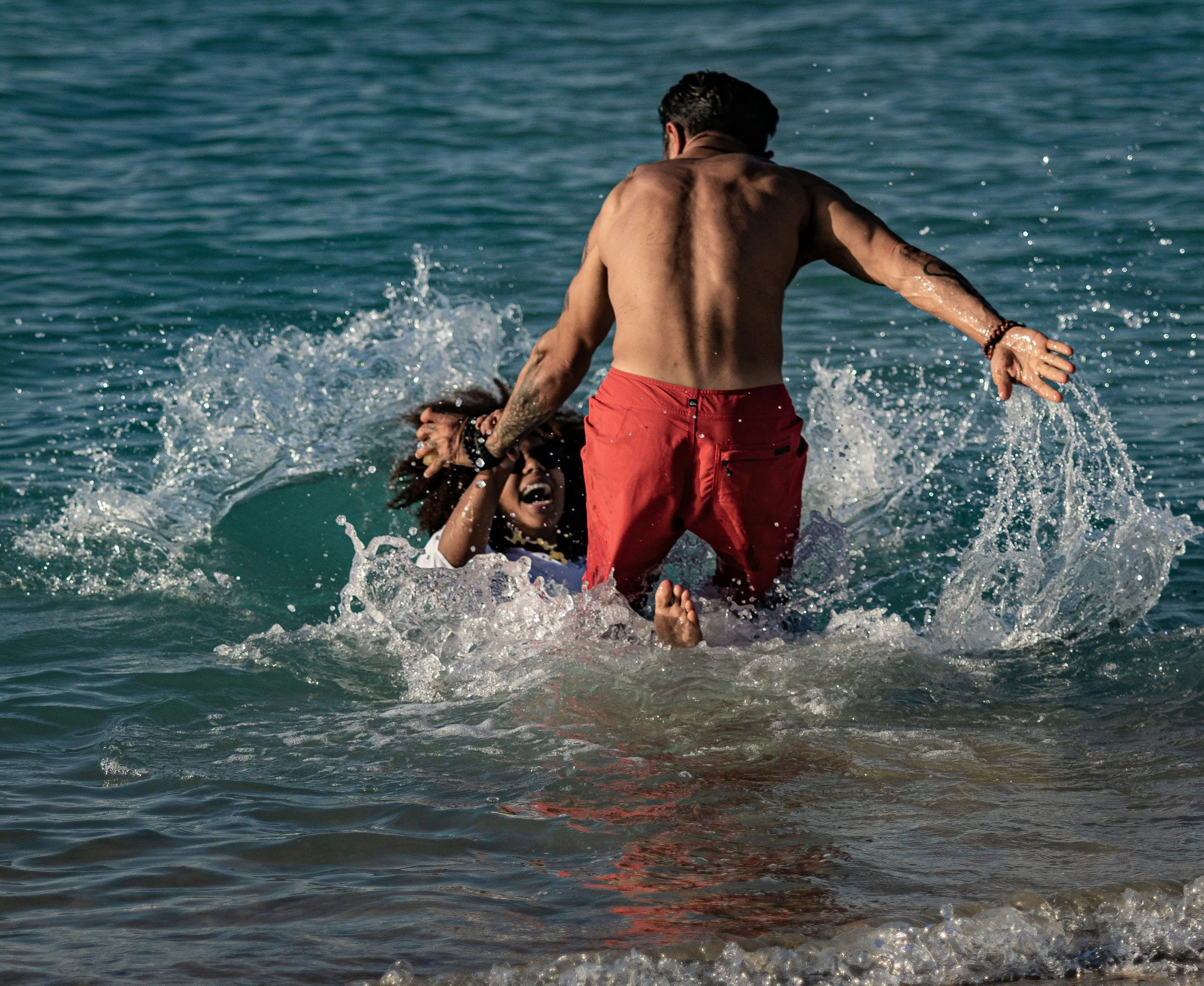 A shirtless man wearing red shorts standing in the ocean, holding a woman under the armpits, and they are both partially submerged in the water.