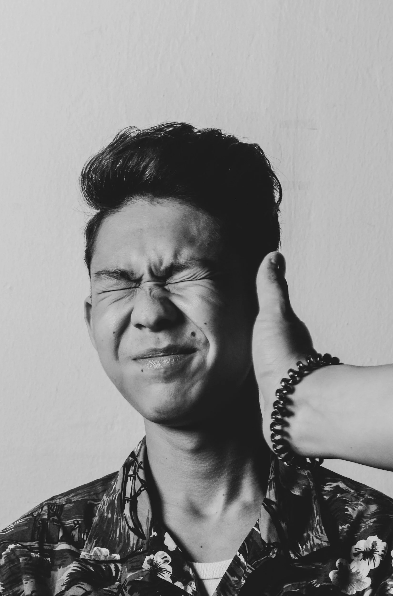 Black and white photo of a young man with closed eyes, smiling, wearing a patterned shirt and a beaded bracelet, with his hand touching his head.