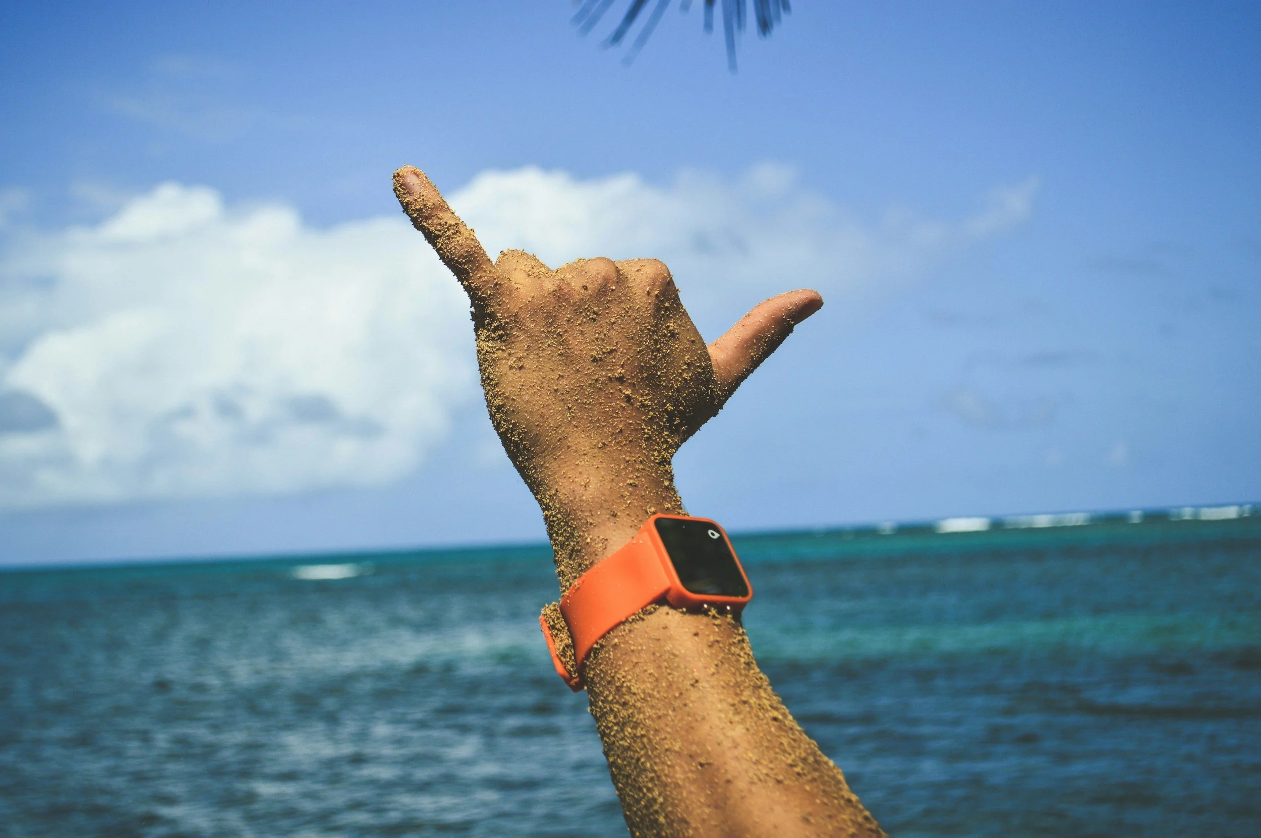 A person's hand with sand on it, wearing an orange wristband and holding a smartphone, is making a shaka sign against a beach and ocean background with blue sky and clouds.