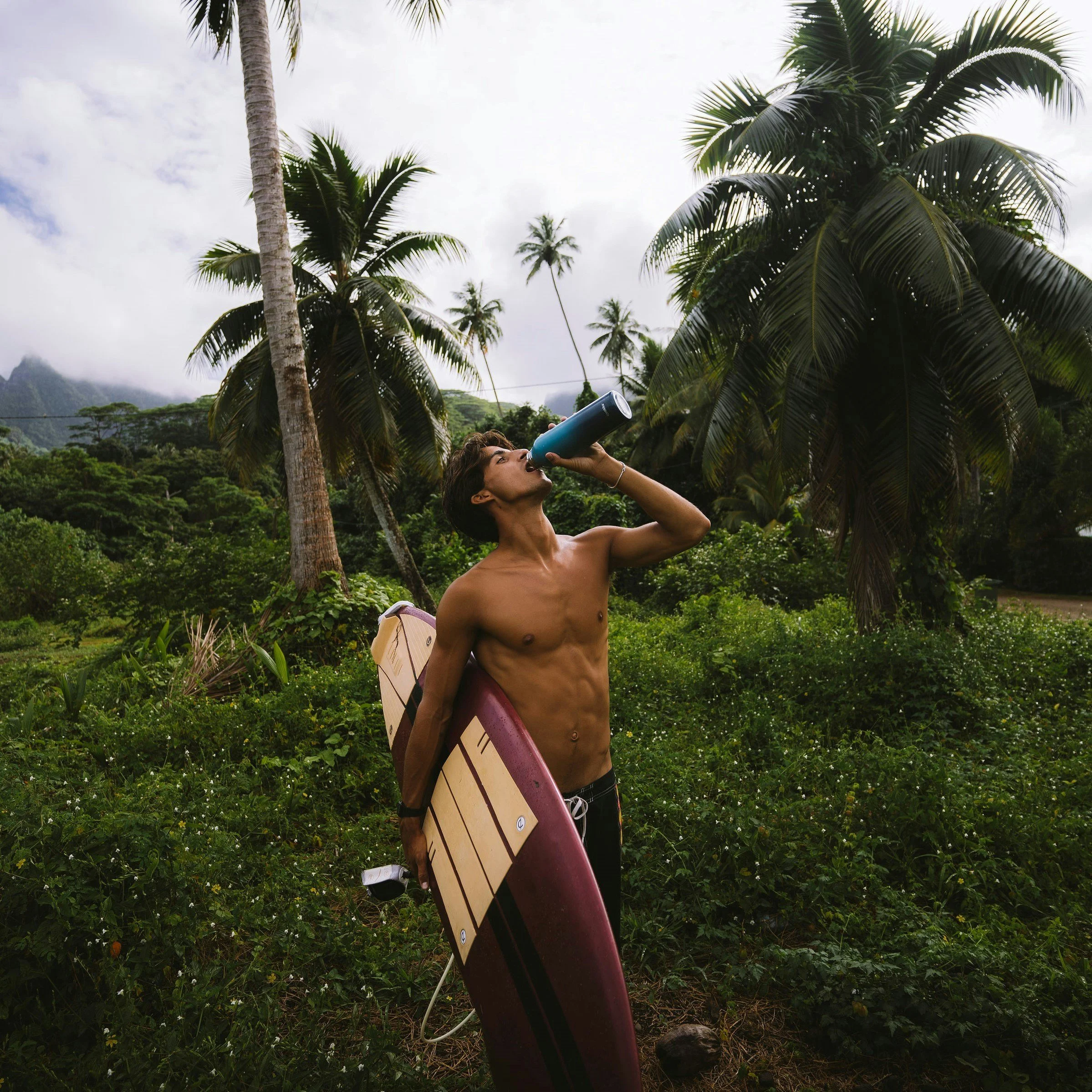 A shirtless man with dark hair and tan skin standing outdoors in a lush green tropical area, holding a blue water bottle to his mouth, with a pink and white surfboard strapped to his left side and a black watch on his left wrist. There are palm trees and dense greenery in the background under a cloudy sky.