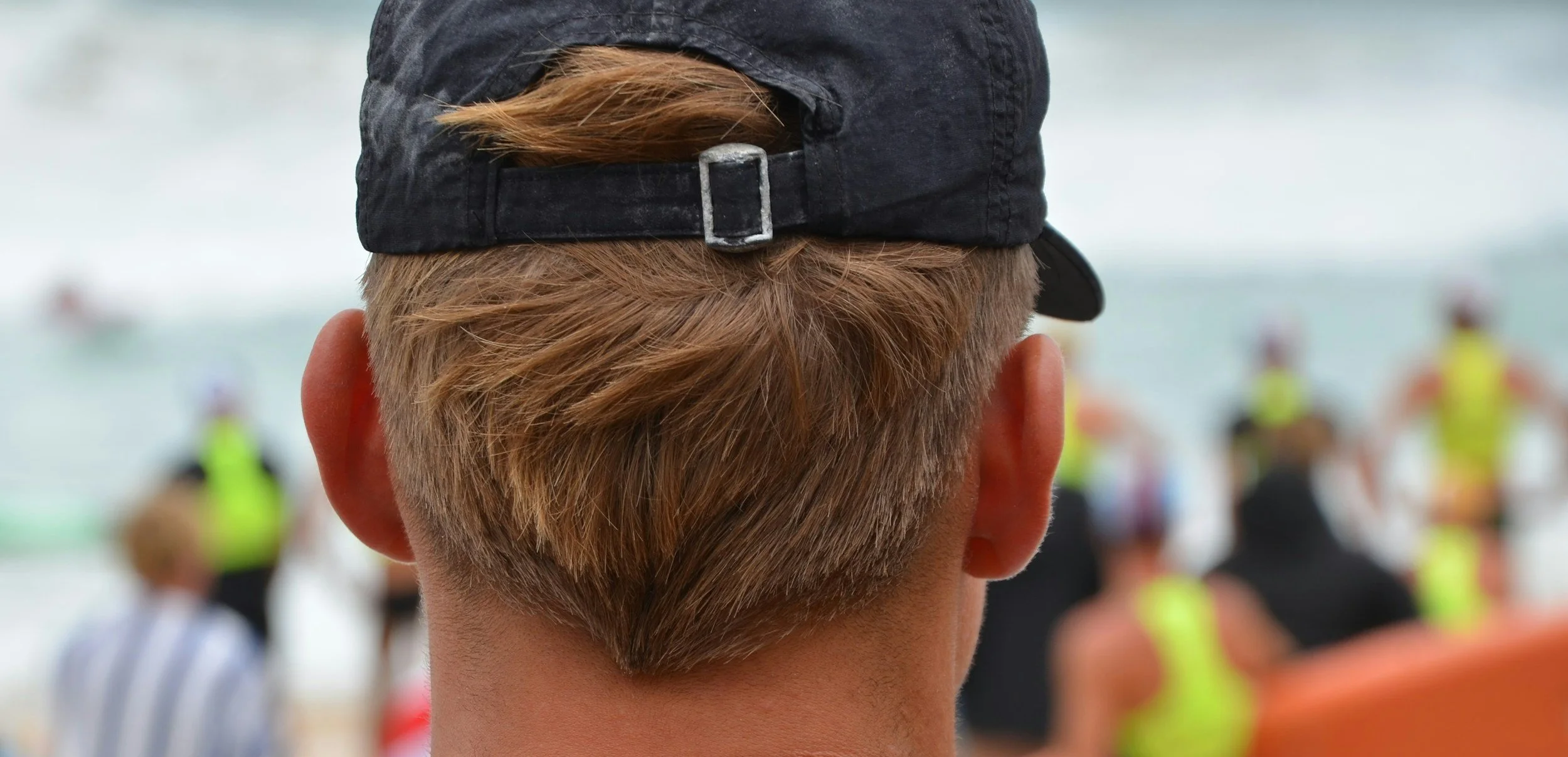 Rear view of a person's head with short, light brown hair, wearing a black baseball cap. The background shows a group of people outdoors near a body of water, some wearing bright clothing, possibly at a beach or outdoor event.