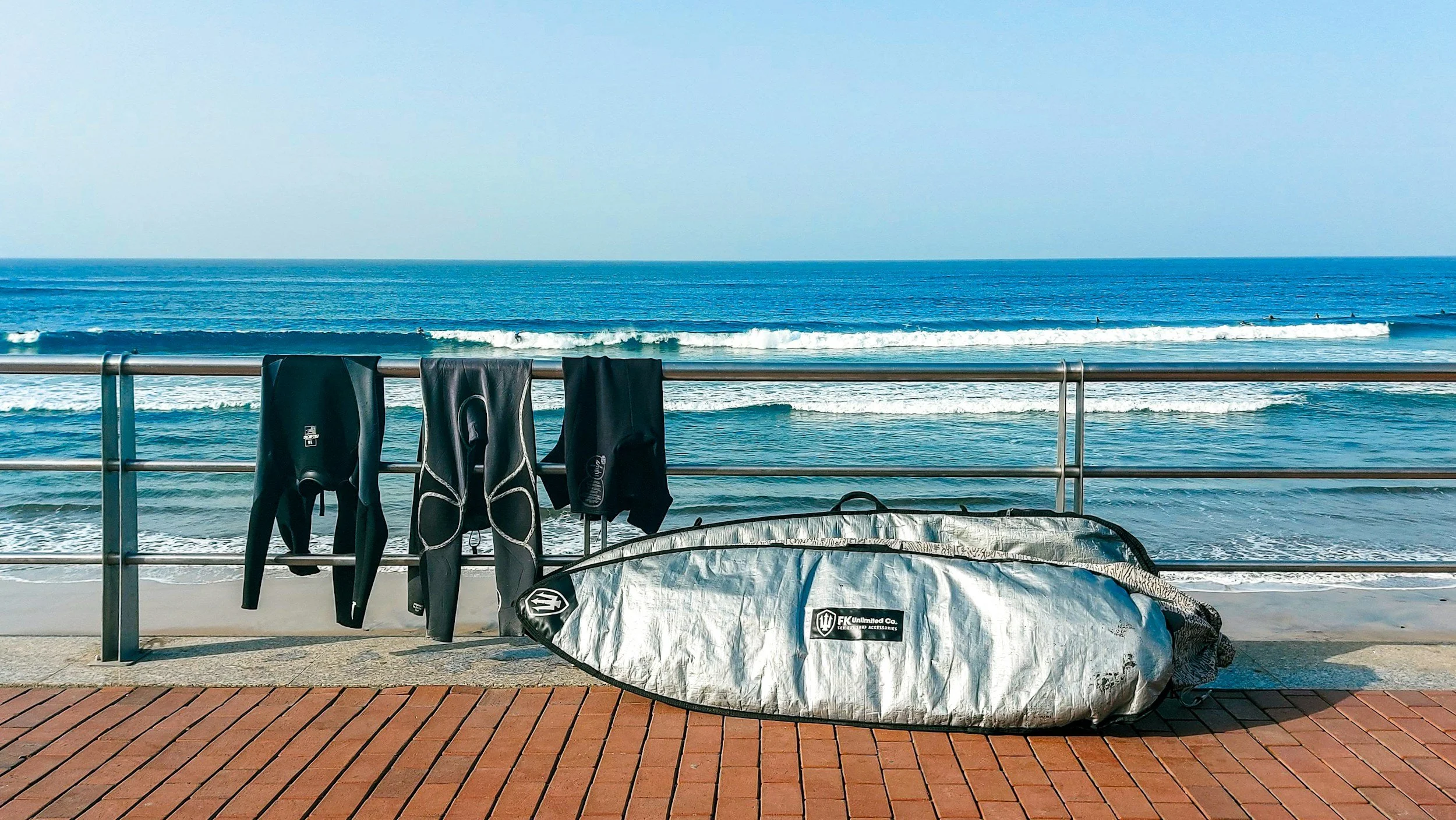 Surfboards and wetsuits hanging on a railing at the beach with waves and a clear sky in the background.