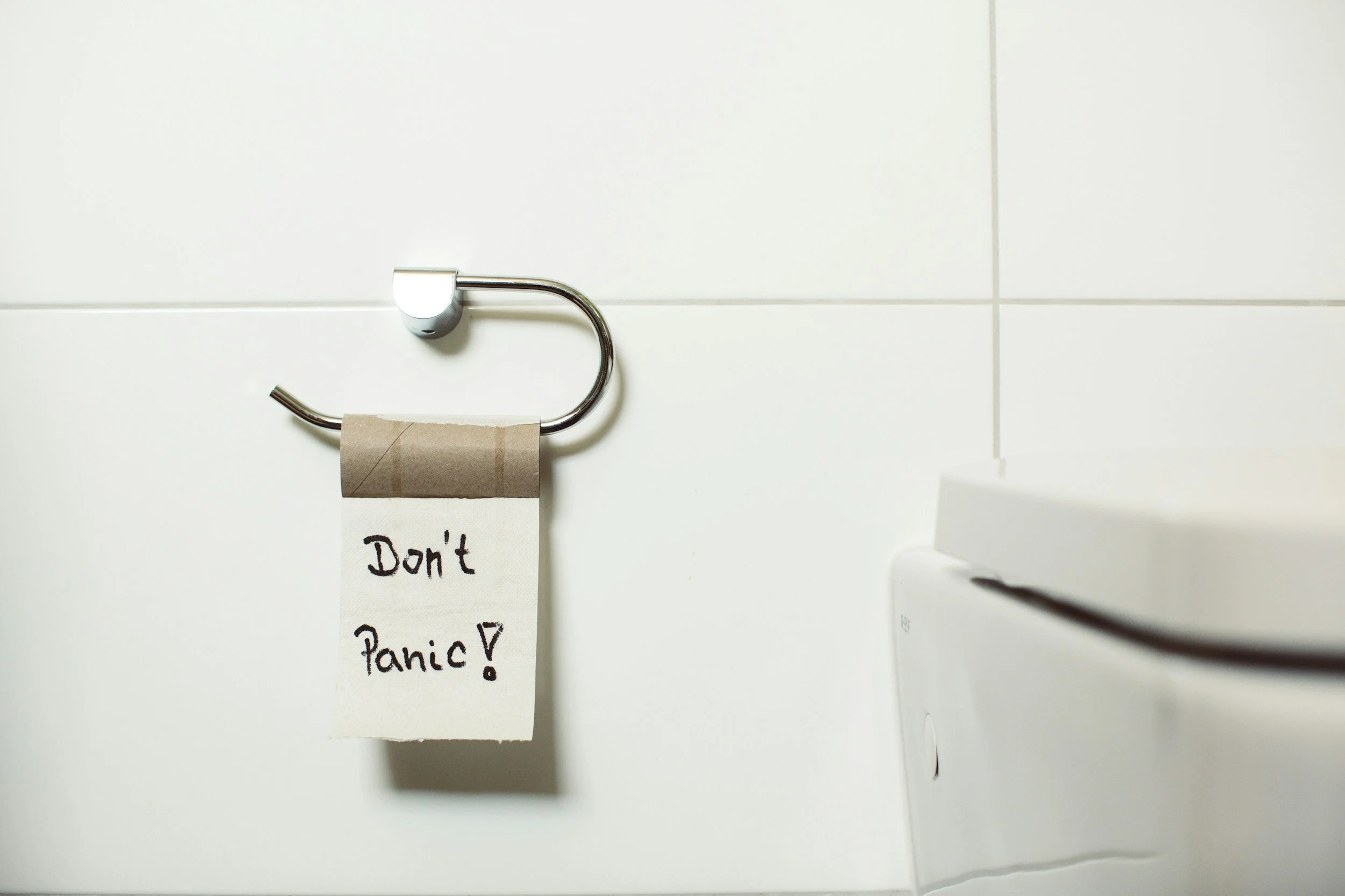 A toilet paper holder with a roll of toilet paper and a note hanging from it that says 'Don't Panic!'