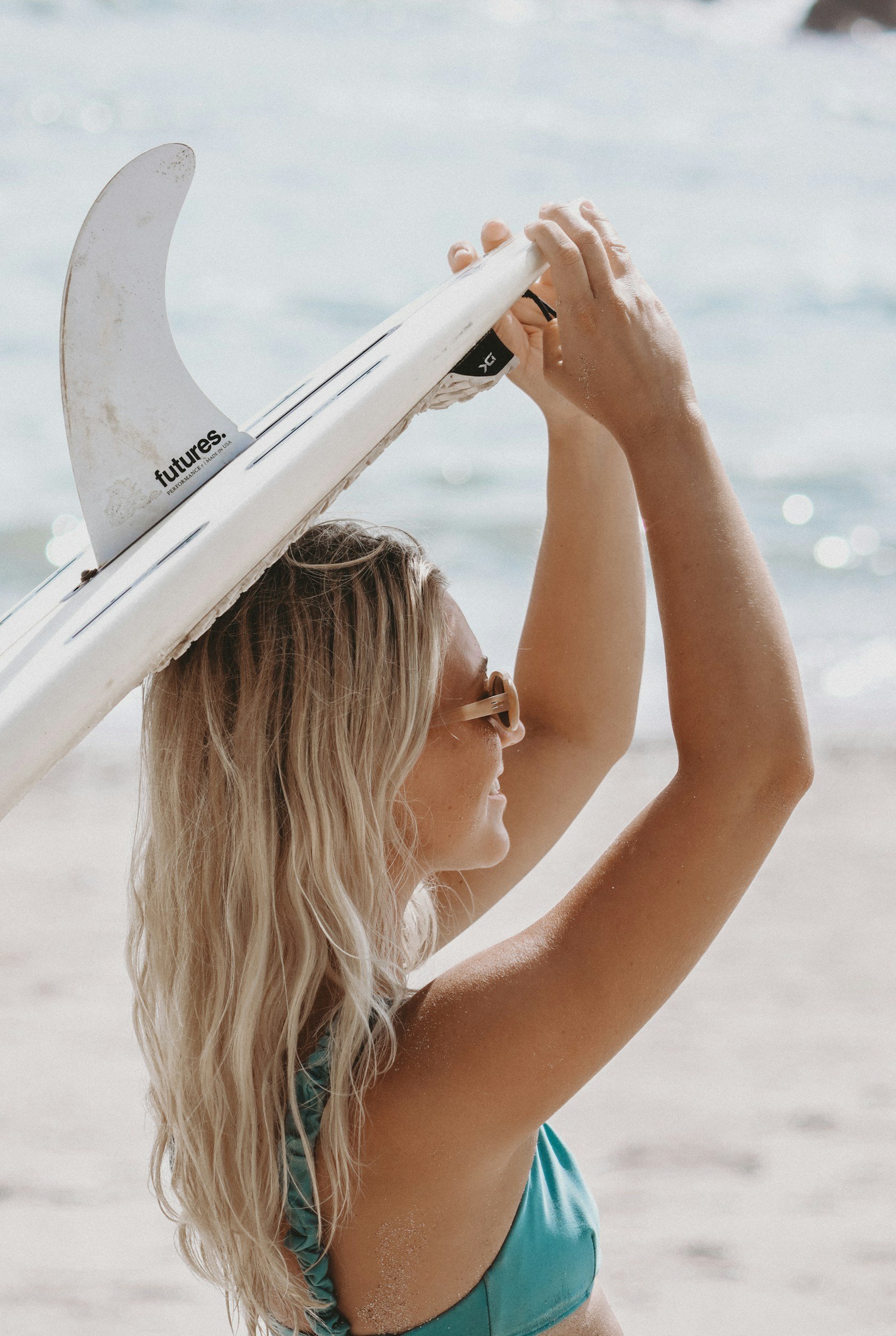A woman with long blonde hair and glasses holding a white surfboard on a beach, with the ocean in the background.