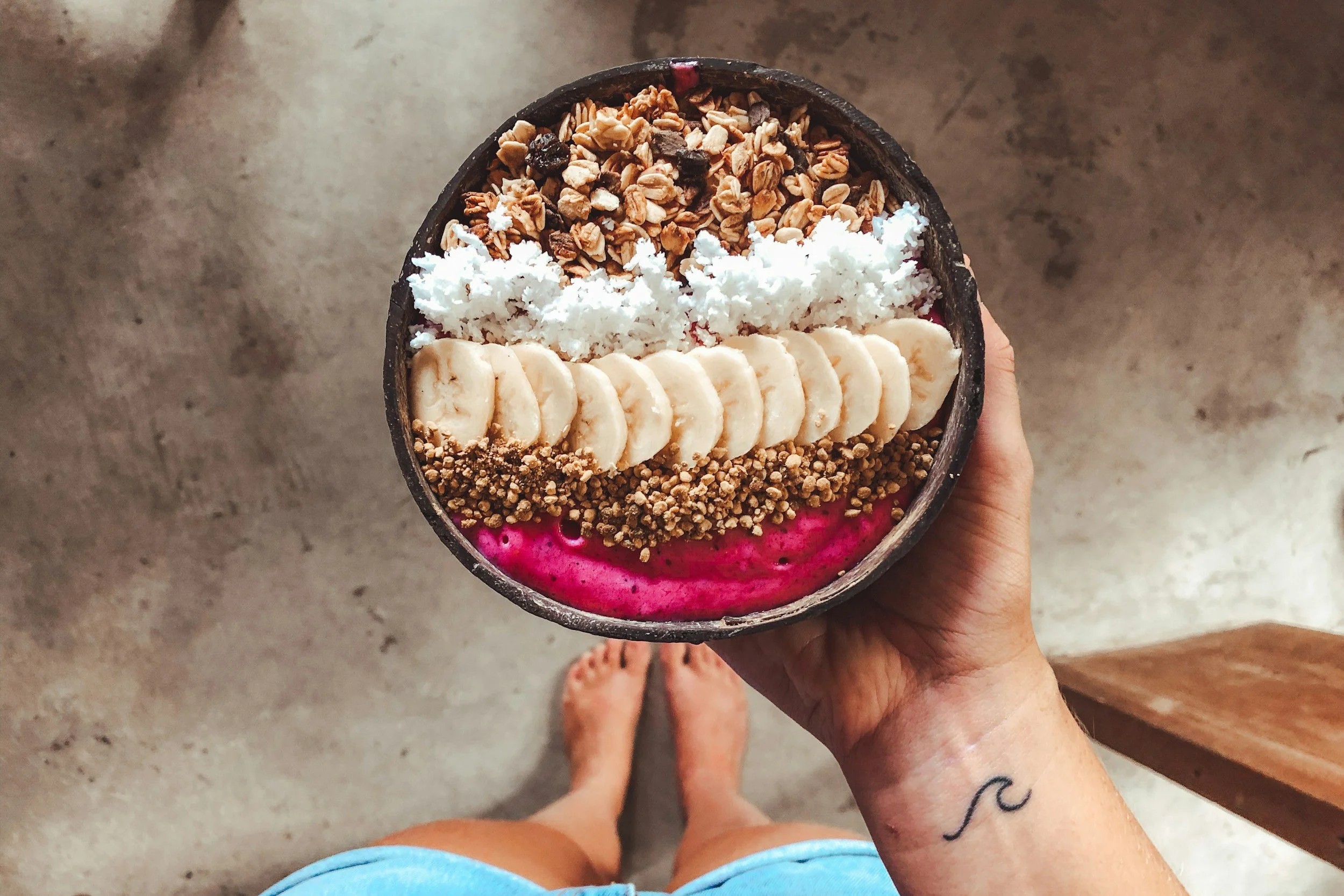 A person holding a bowl of colorful smoothie with sliced bananas, shredded coconut, granola, and a pink smoothie base.