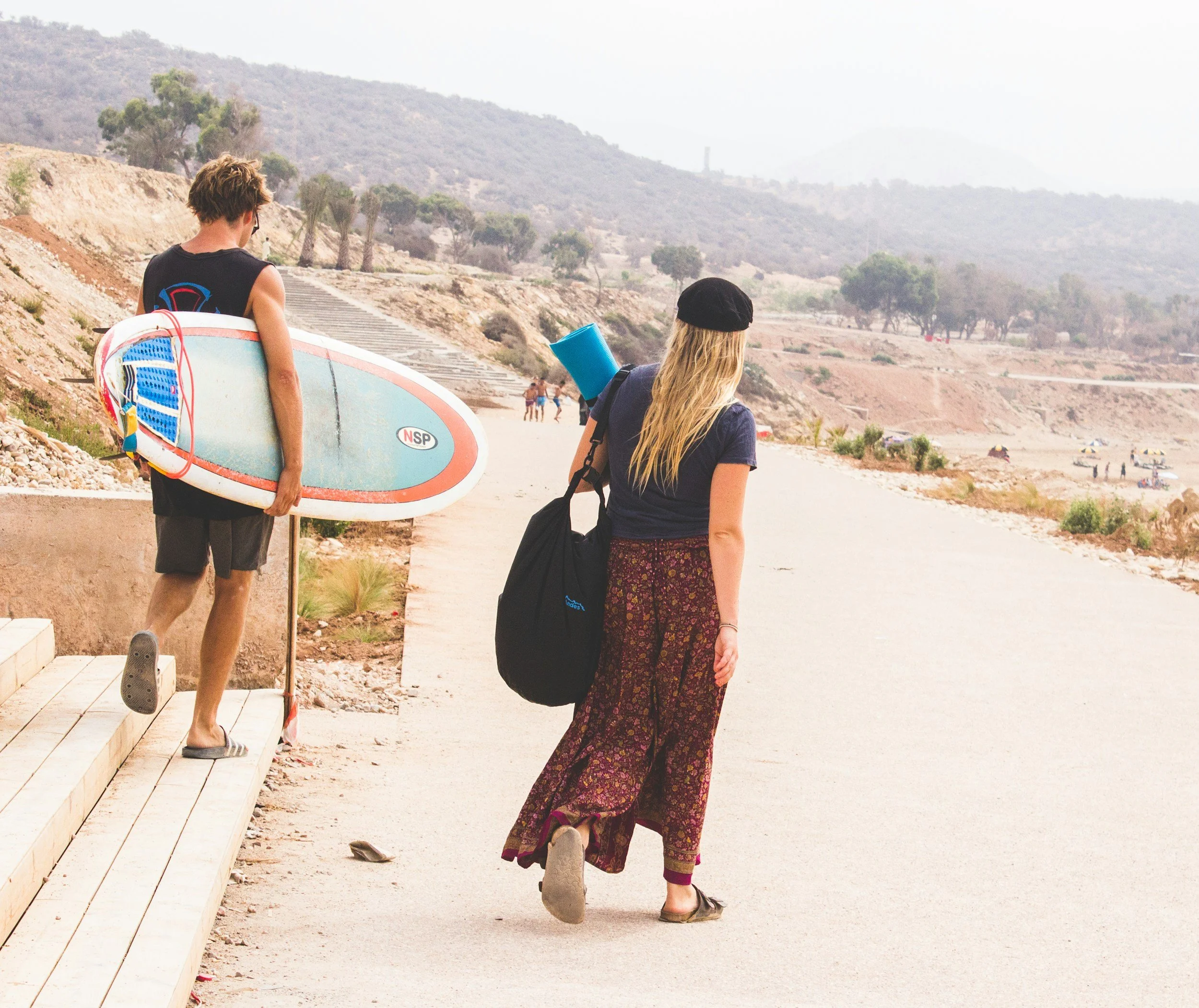 Two people walking on a beach with a mountainous landscape in the background. One person, with blonde hair, is wearing a black hat, navy blue top, long patterned skirt, and carrying a black bag. The other person, with short hair, is holding a surfboard, wearing shorts and a sleeveless top.