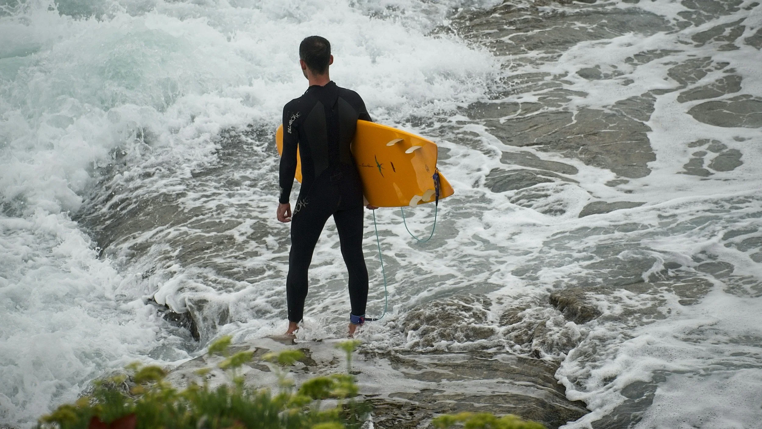 A man in a wetsuit holding a yellow surfboard, standing at the edge of the water on a rocky shoreline.