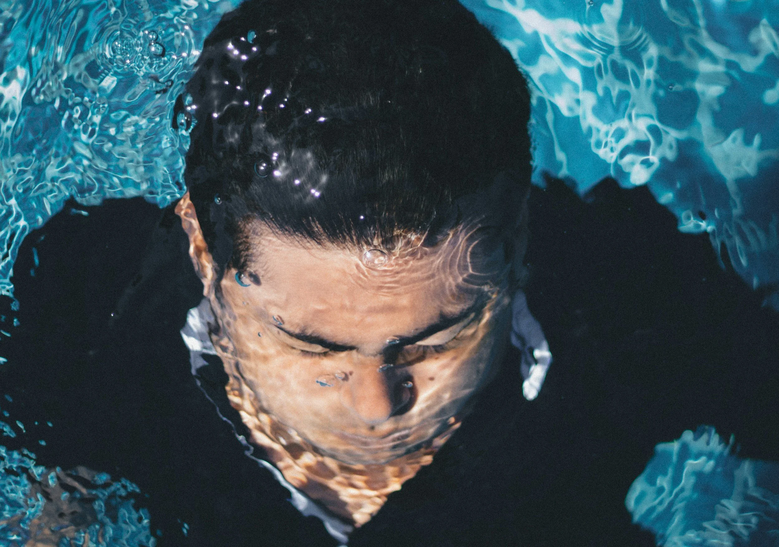 A person swimming underwater with their head submerged, surrounded by blue water, with bubbles forming around their face.