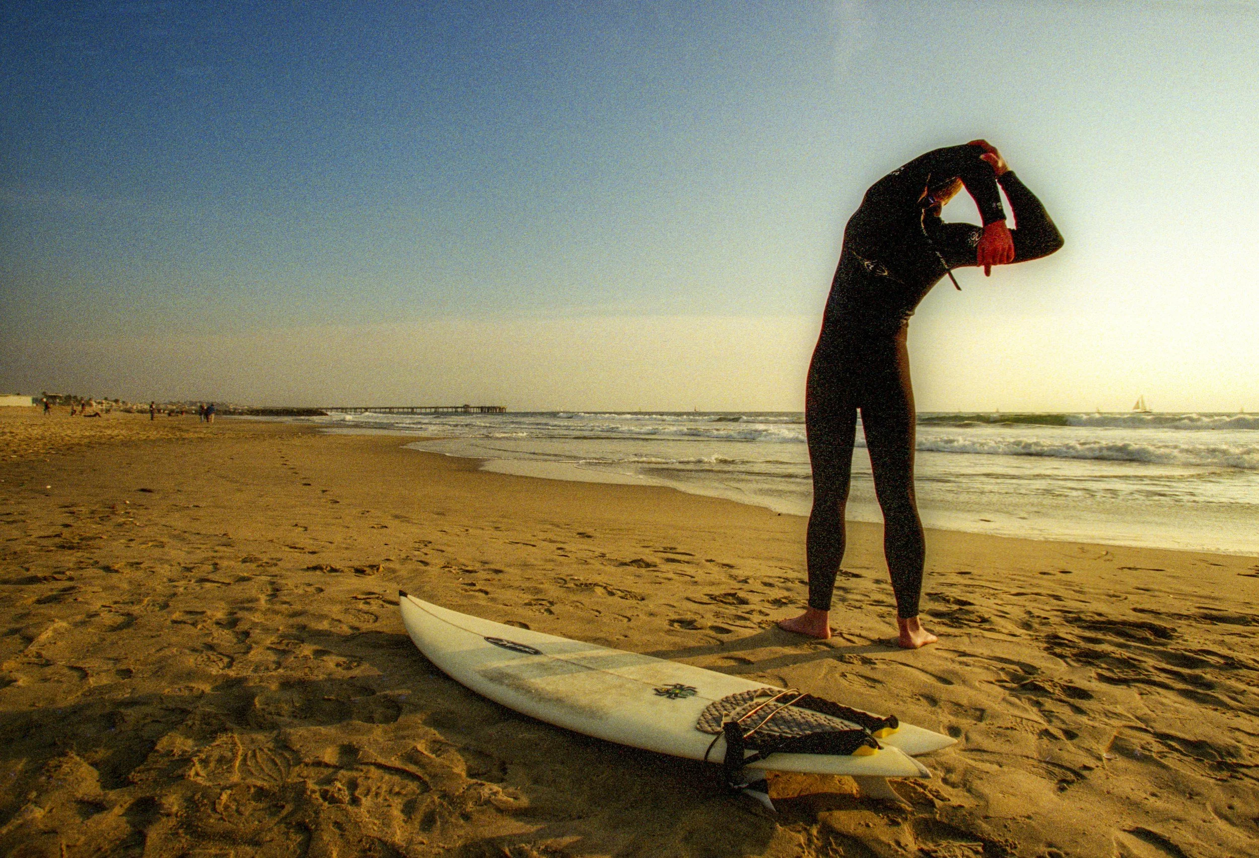 Person standing on a sandy beach at sunset, holding their head, with a stand-up paddleboard and paddle in the foreground, and a pier in the distance.
