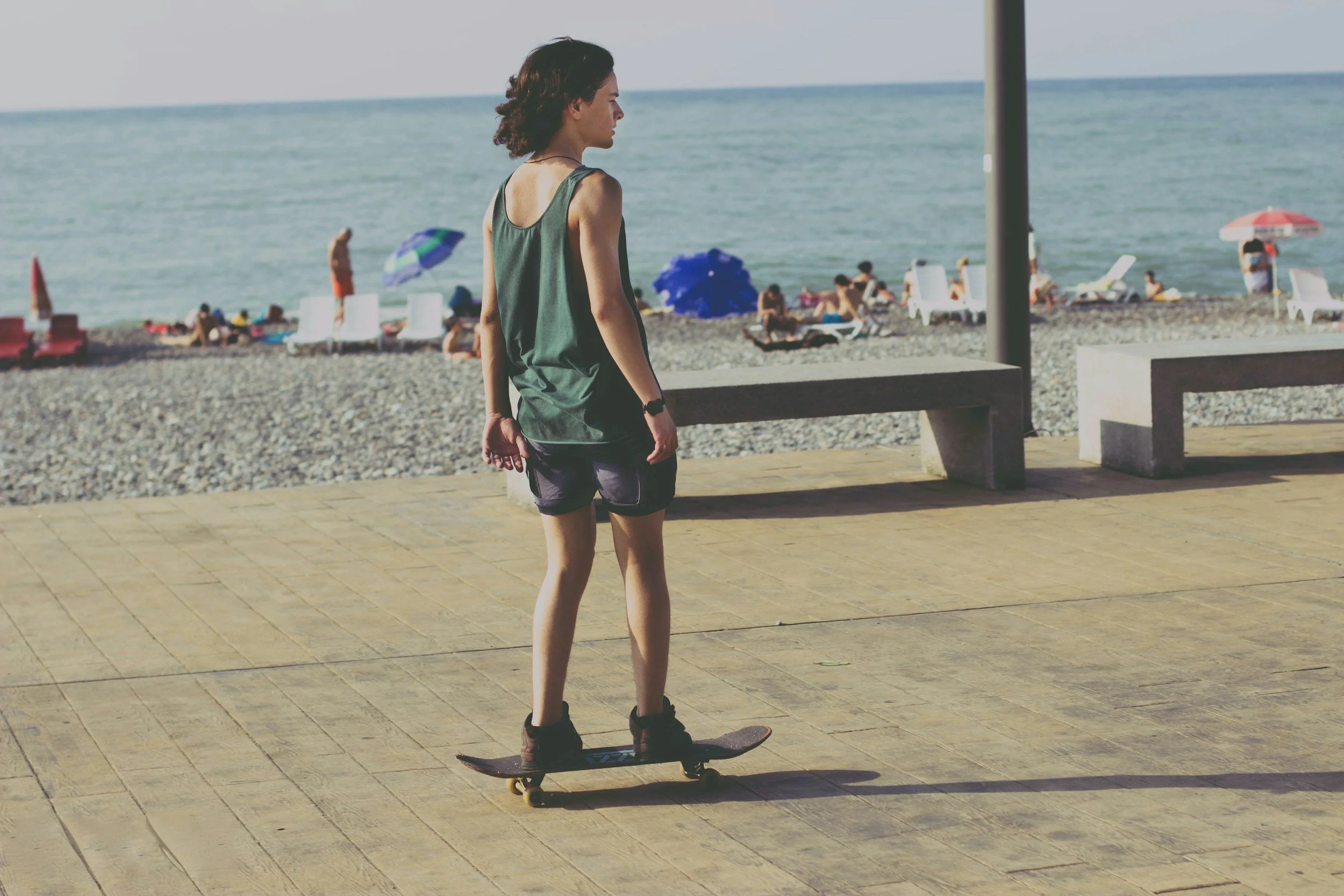 Young person with curly hair skateboarding near a beach, with umbrellas and people relaxing on the shore in the background.