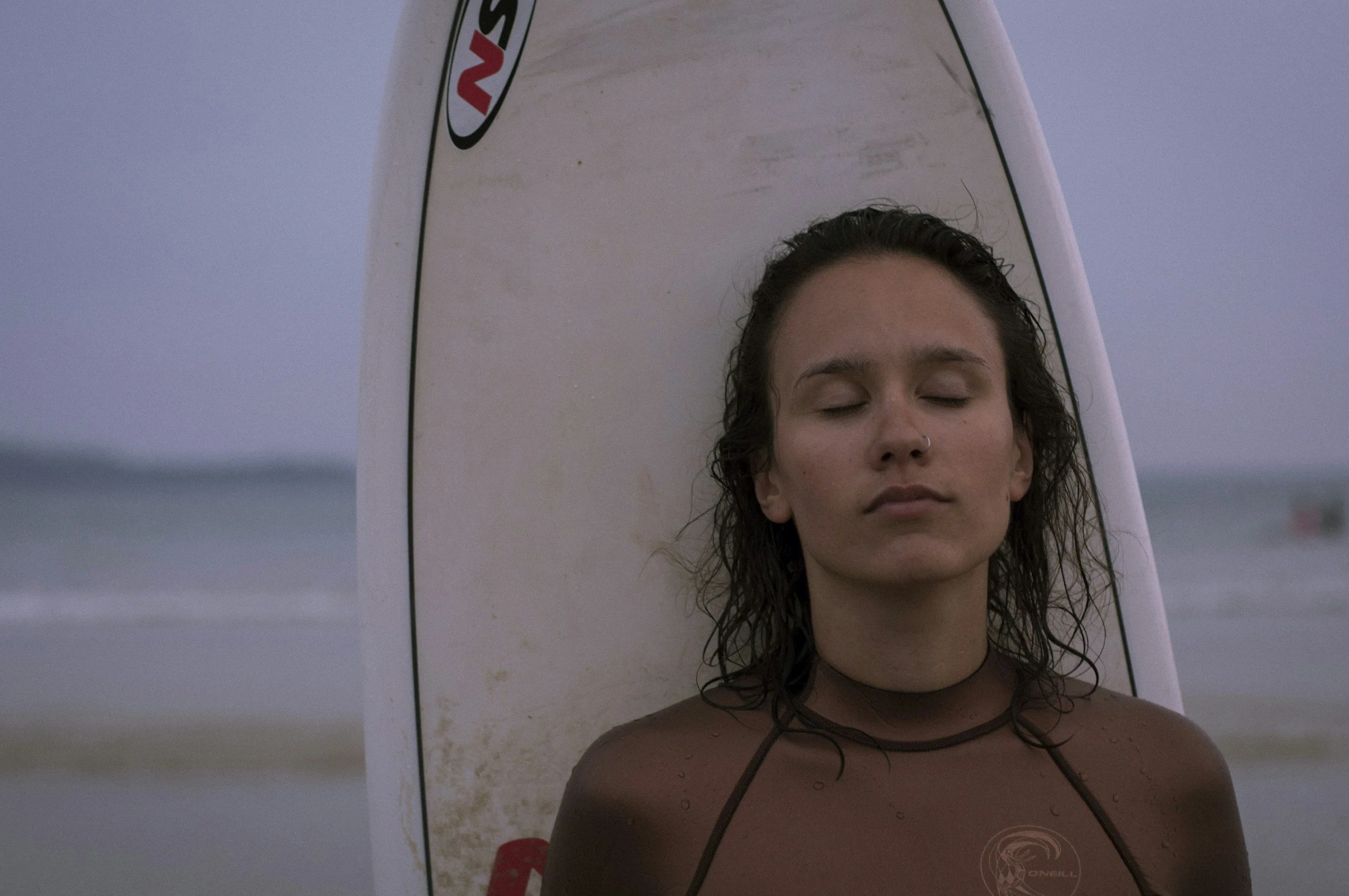 A woman with wet, curly hair and closed eyes holding a surfboard behind her head at the beach.
