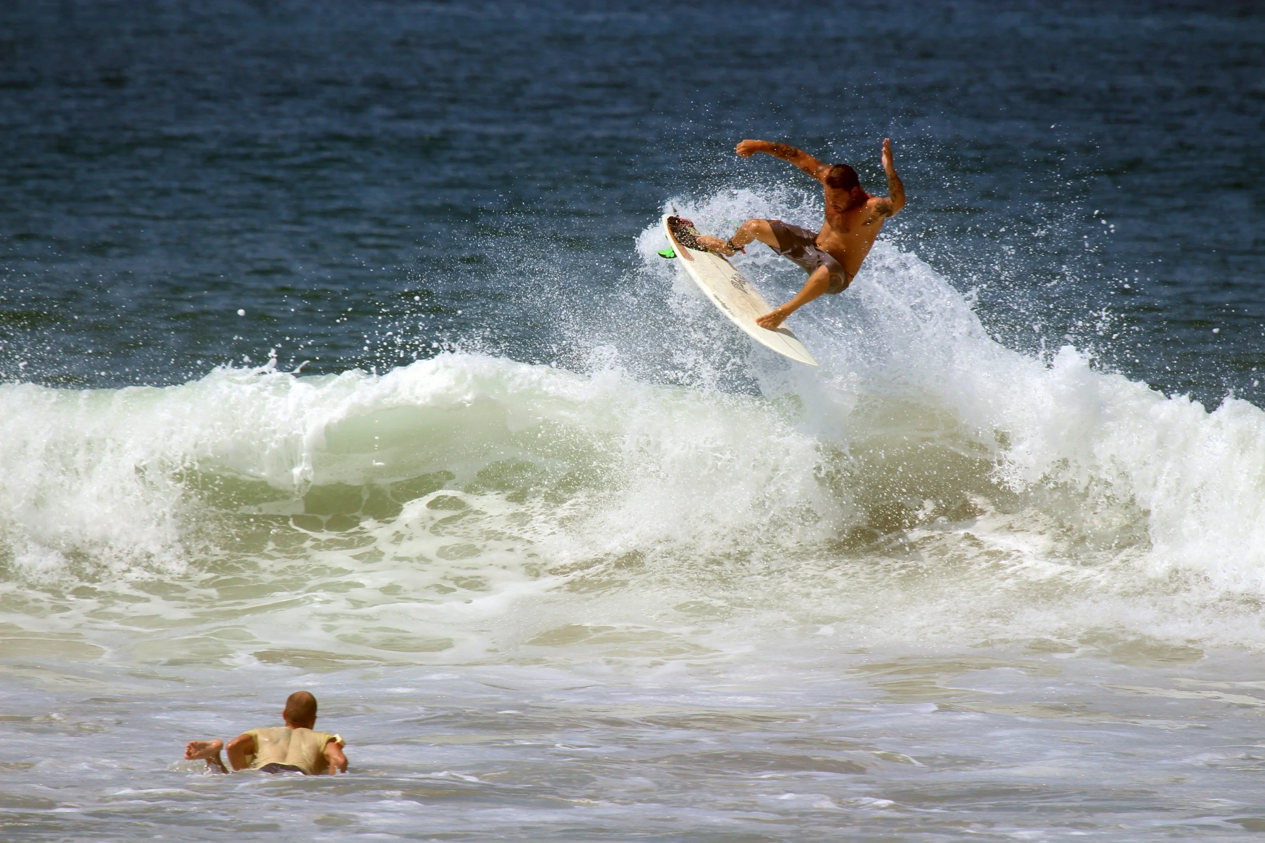 A man with tattoos surfing on a wave in the ocean, with a person in the foreground in shallow water.