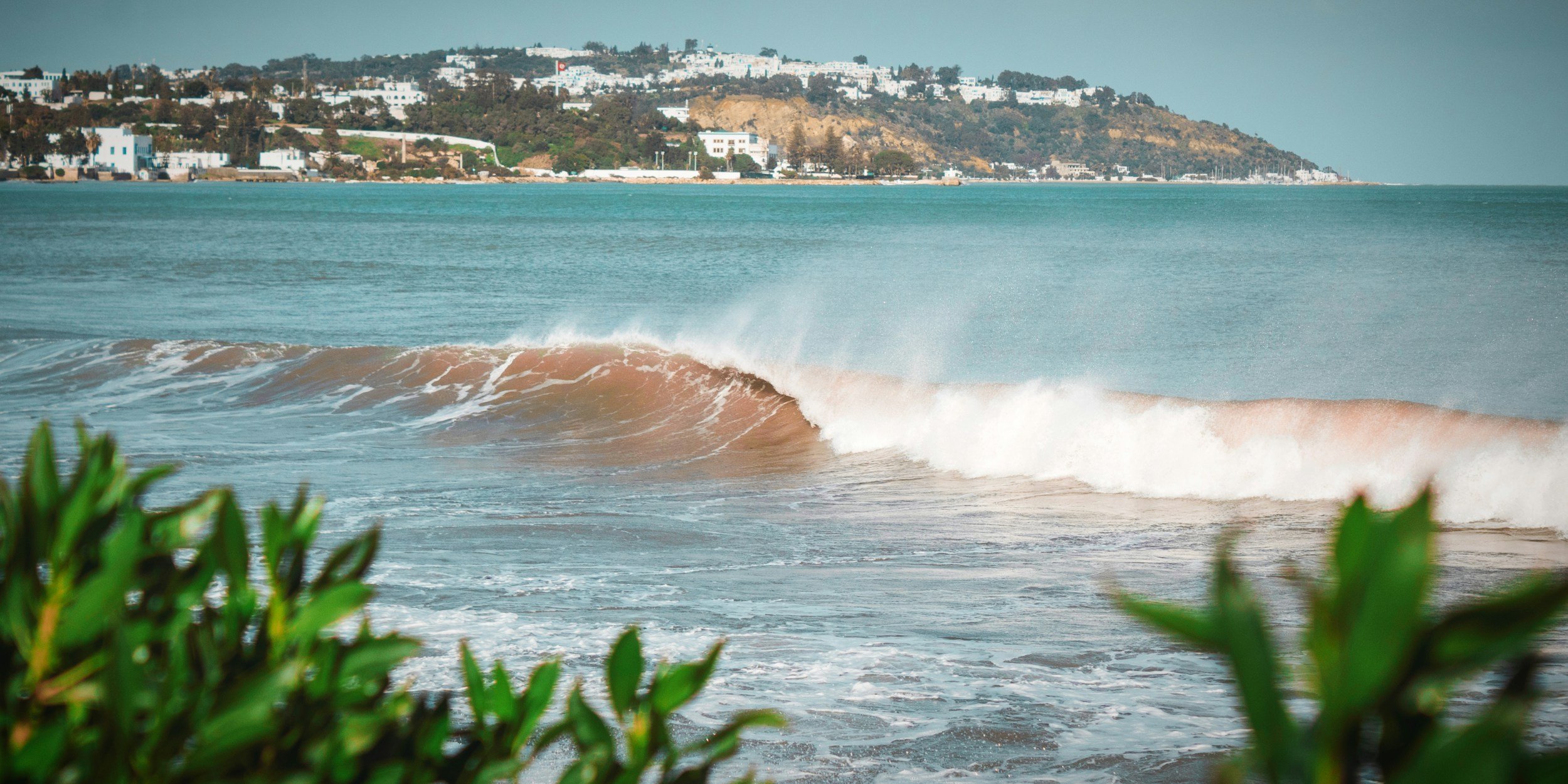 Ocean waves crashing on the beach with a hillside town in the background, featuring white buildings and some greenery.