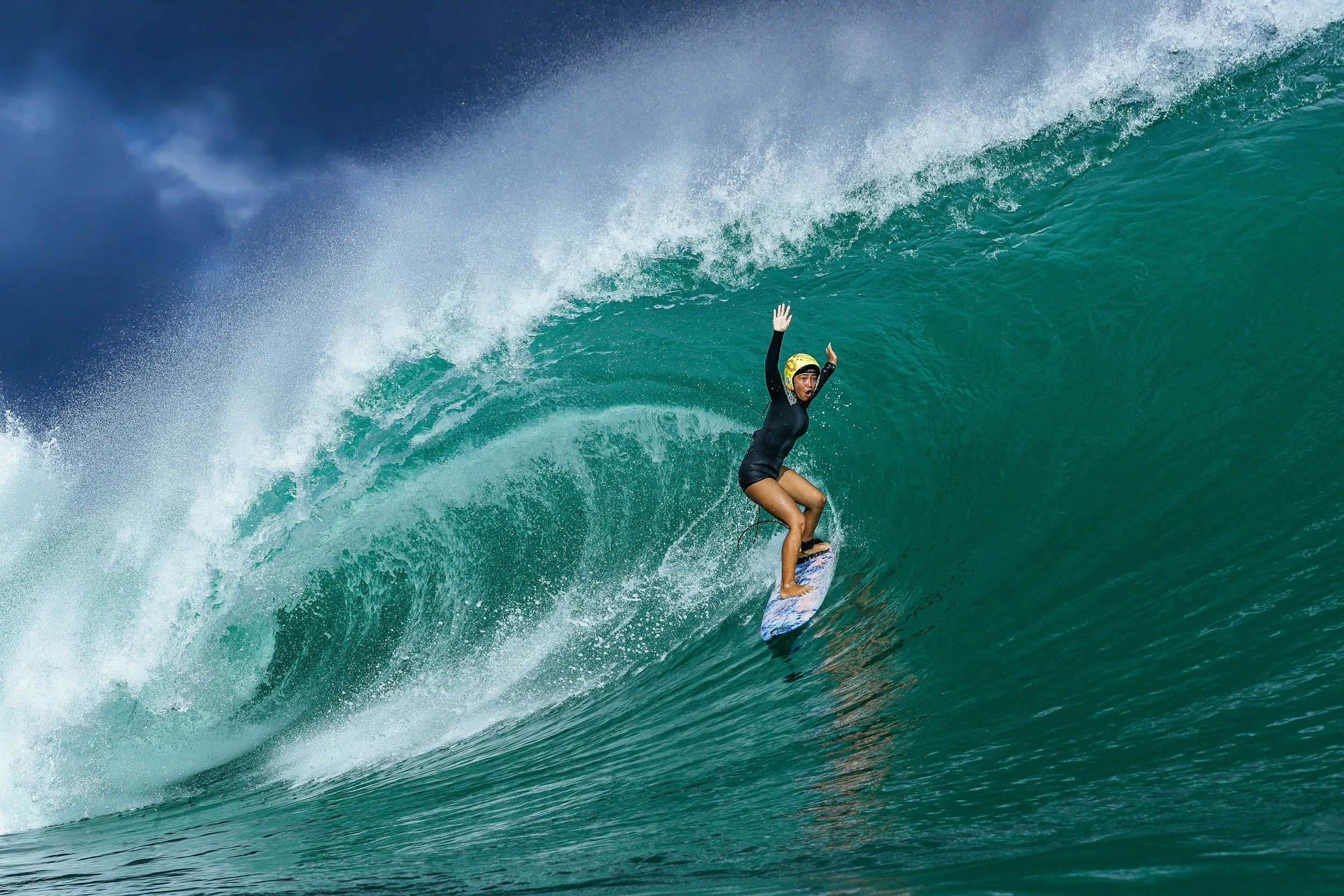 A woman surfing on a large green wave, wearing a black wetsuit, yellow helmet, and standing on a surfboard with her arms raised.