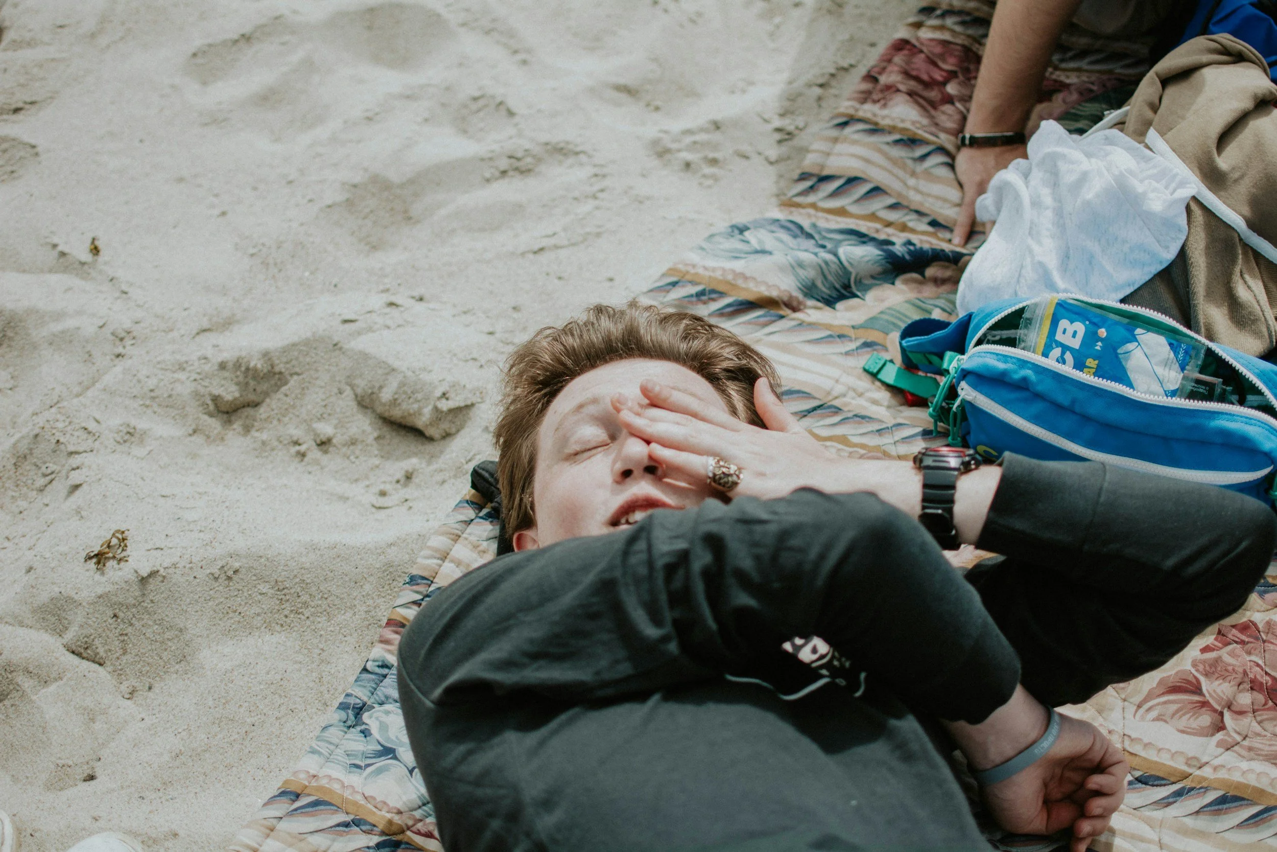 Person lying on a blanket on the sandy beach, covering their eyes with one hand, sick on surf trip