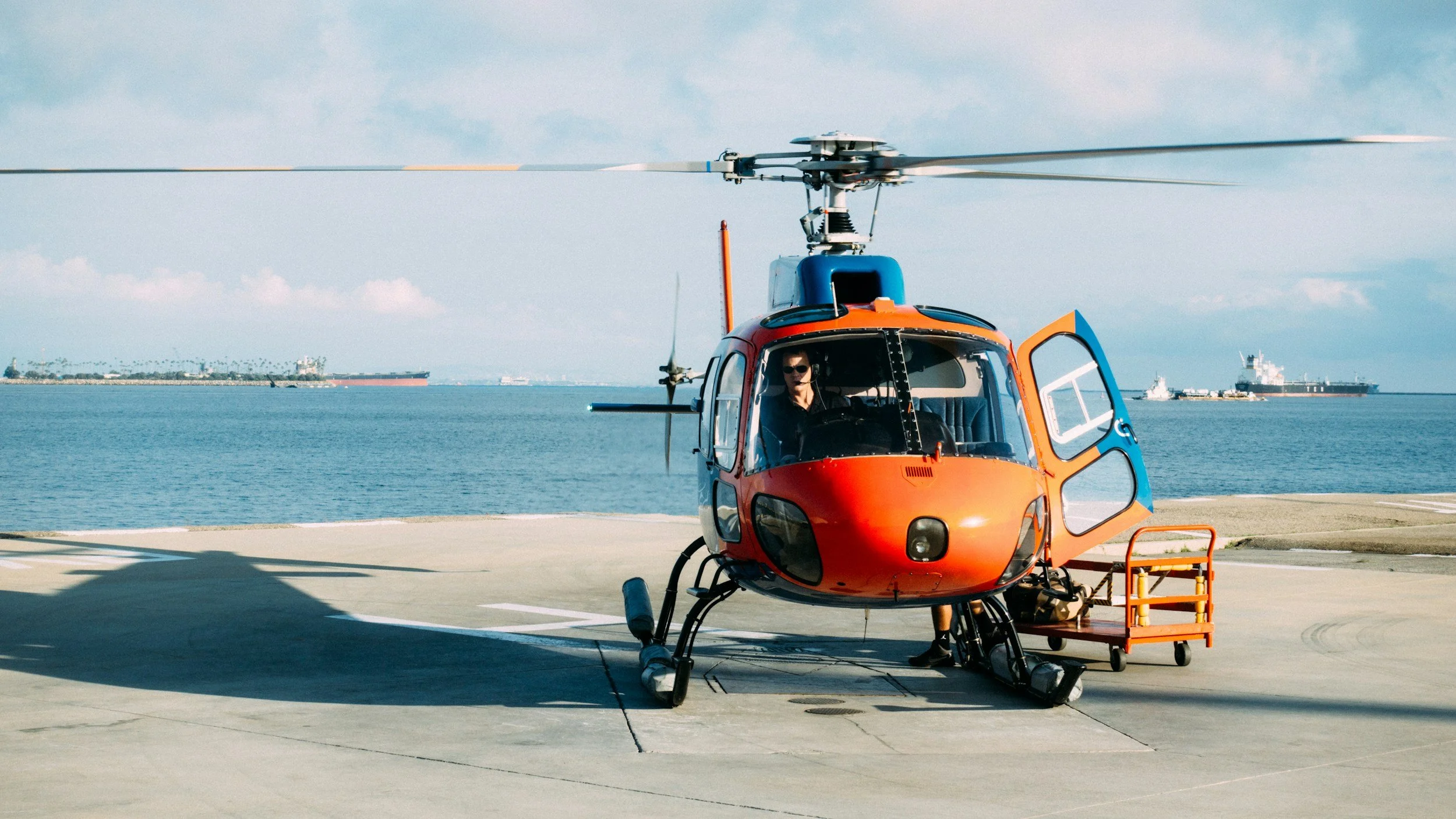 A helicopter parked on a helipad near the water with ships in the background, and a person seated inside the helicopter.