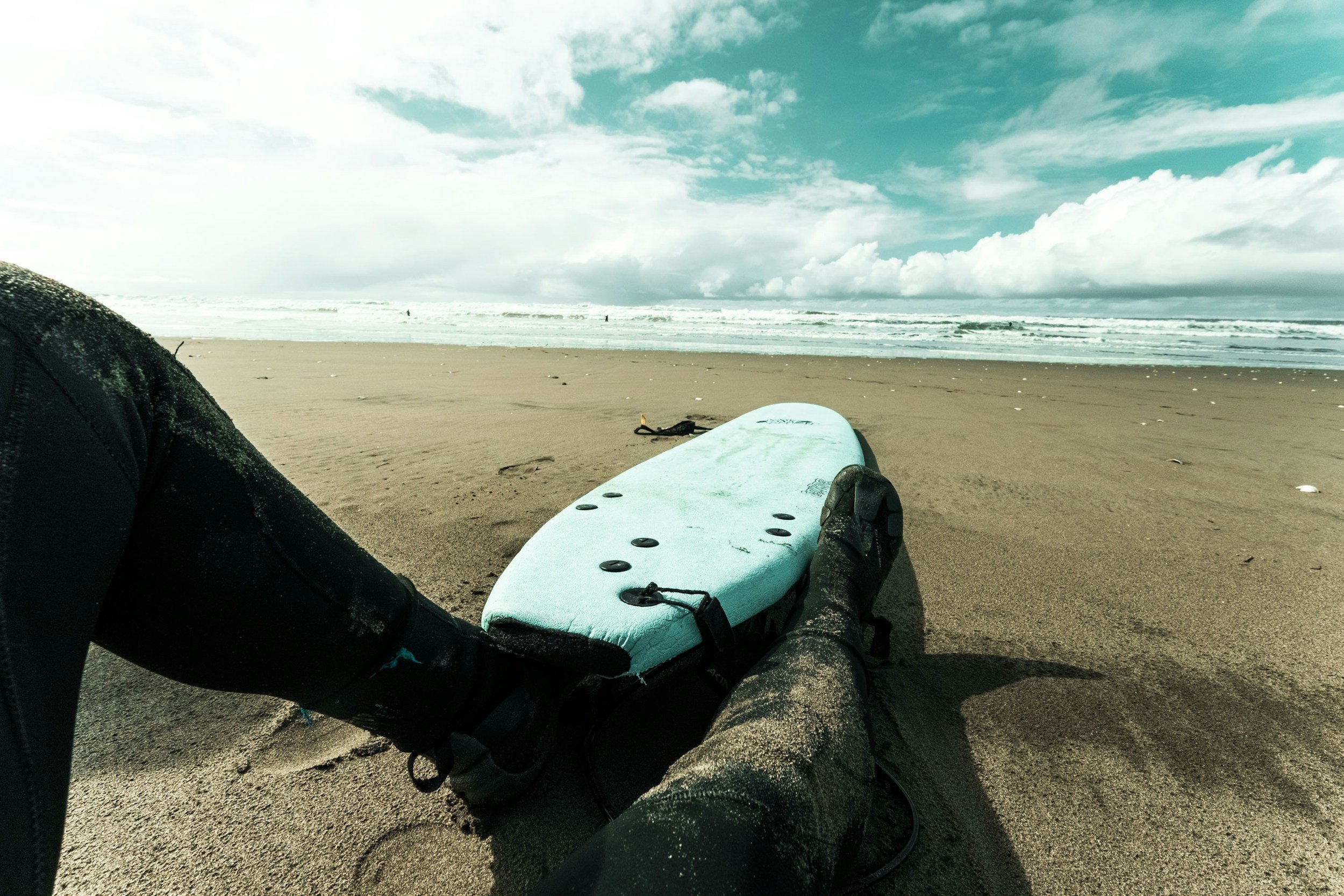 Person sitting on sandy beach with surfboard, experiencing toe cramps likely related to dehyration.