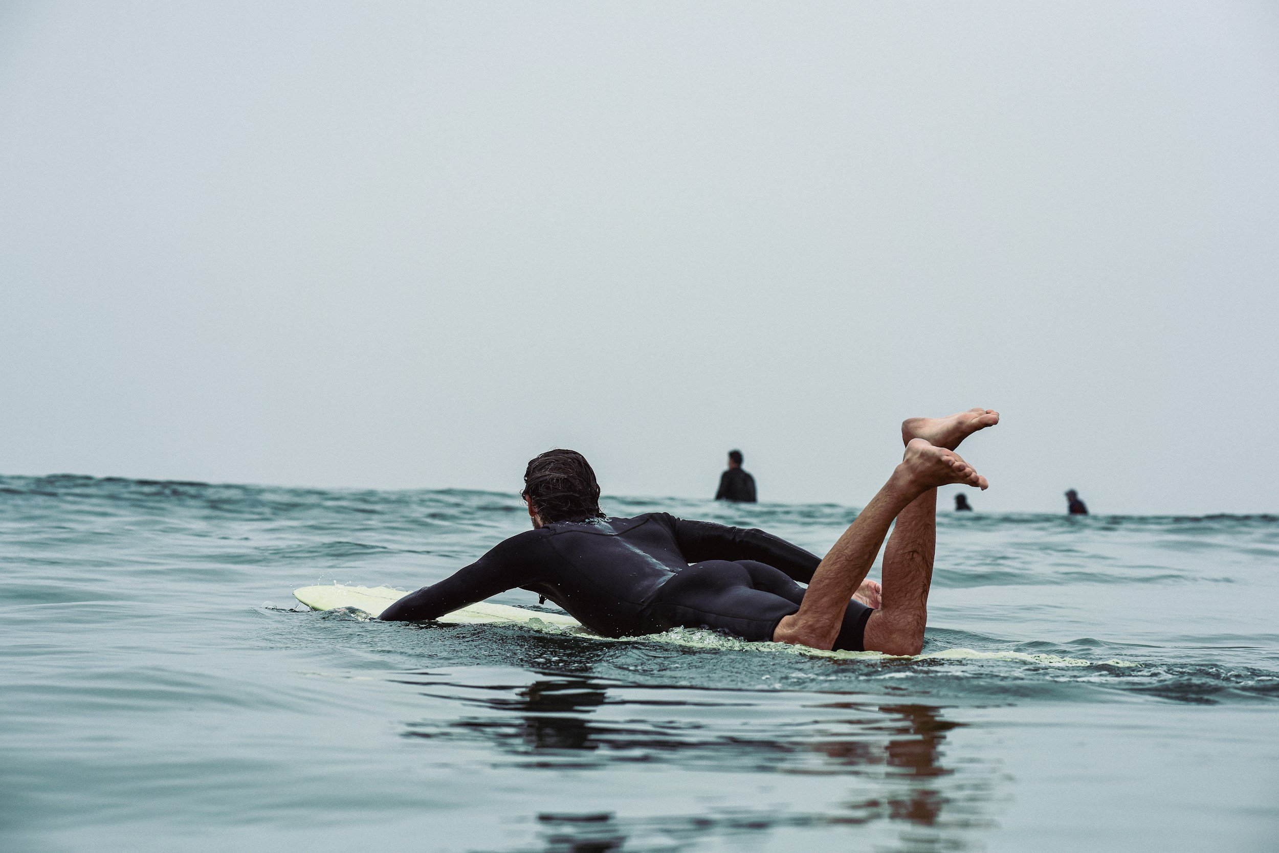 A person lying on a surfboard in the water, with others in the background, and a cloudy sky overhead.  GERD and reflux from surfing, laying flat